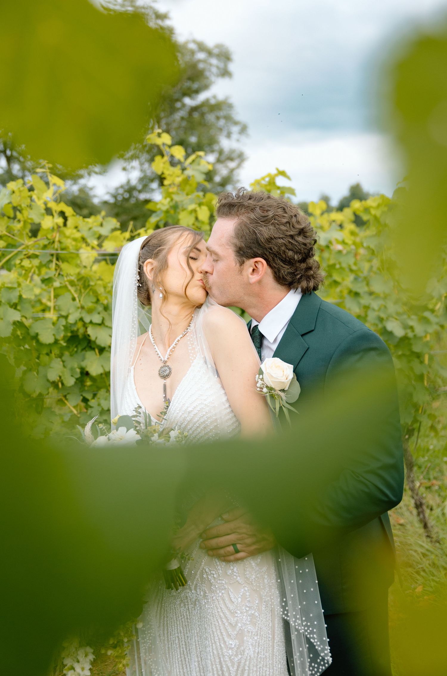 Bride and groom sharing a romantic kiss surrounded by lush green vineyard vines on their wedding day.