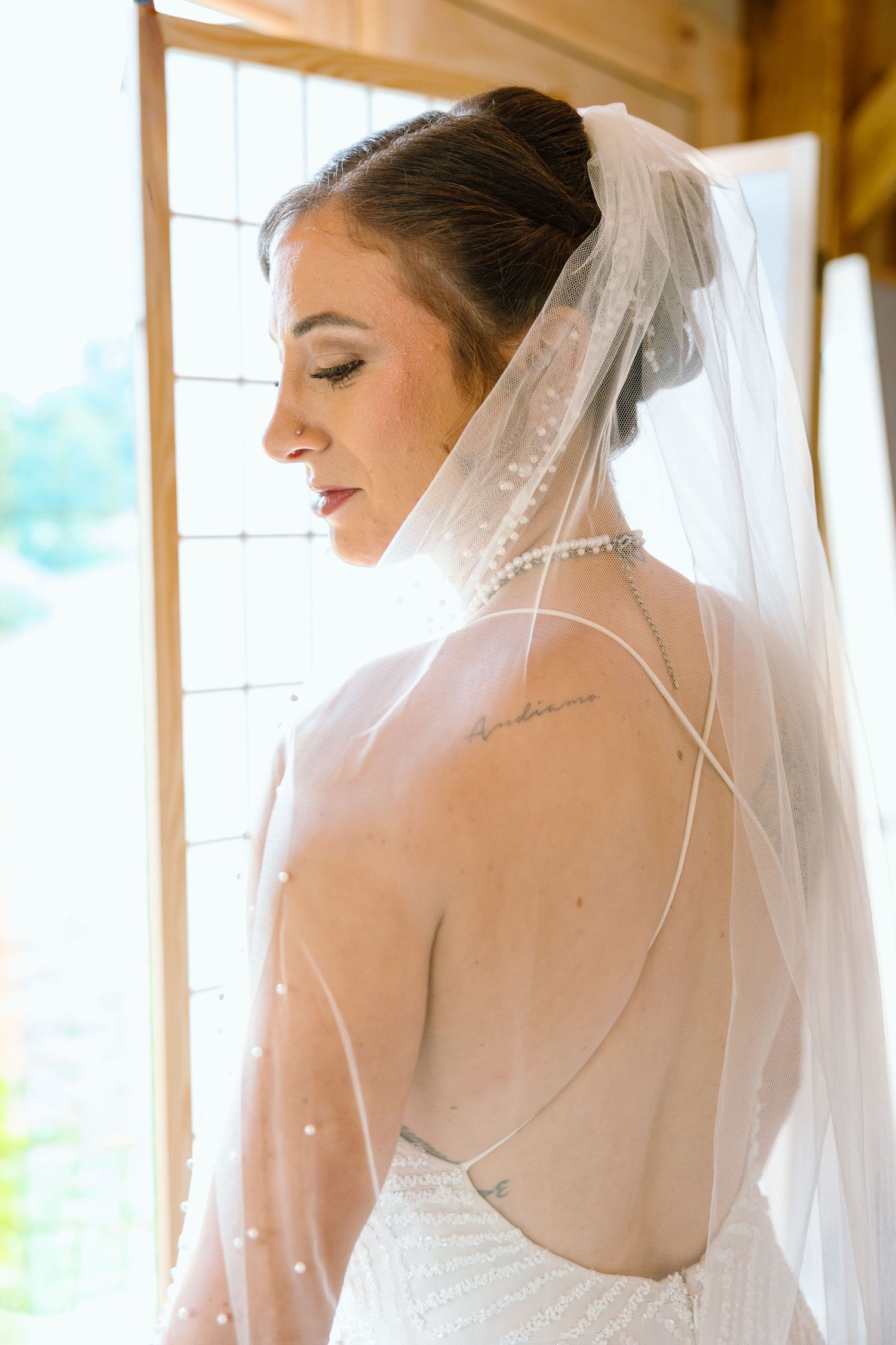 Bride in backless wedding dress and pearl-trimmed veil gazes down by a sunlit window, showcasing elegant updo and shoulder tattoo.