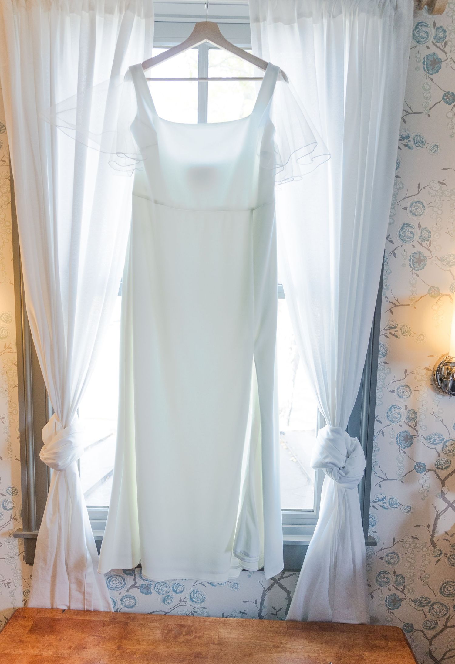 White bridesmaid dresses hanging by a window with sheer curtains and floral wallpaper.
