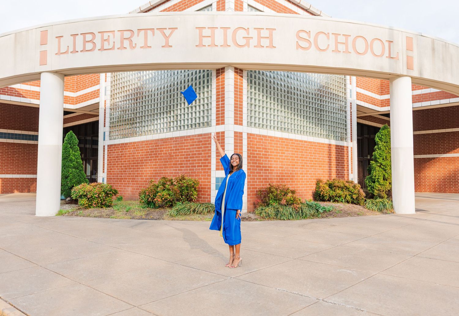 Graduate in bright blue regalia poses triumphantly beneath Liberty High School sign.