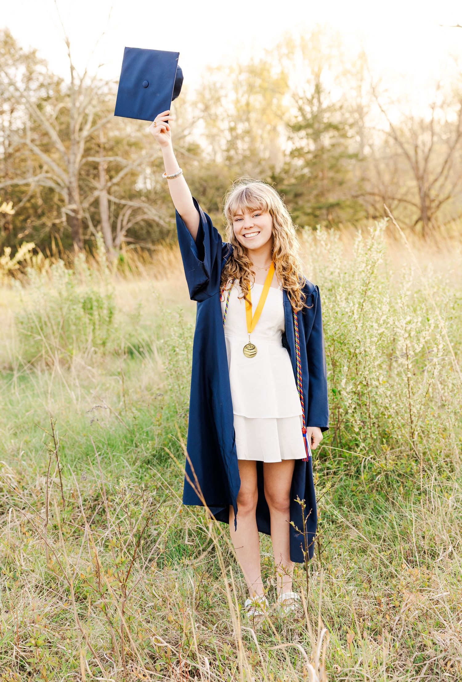 Graduate in white dress and blue cap celebrating in a sunlit field.