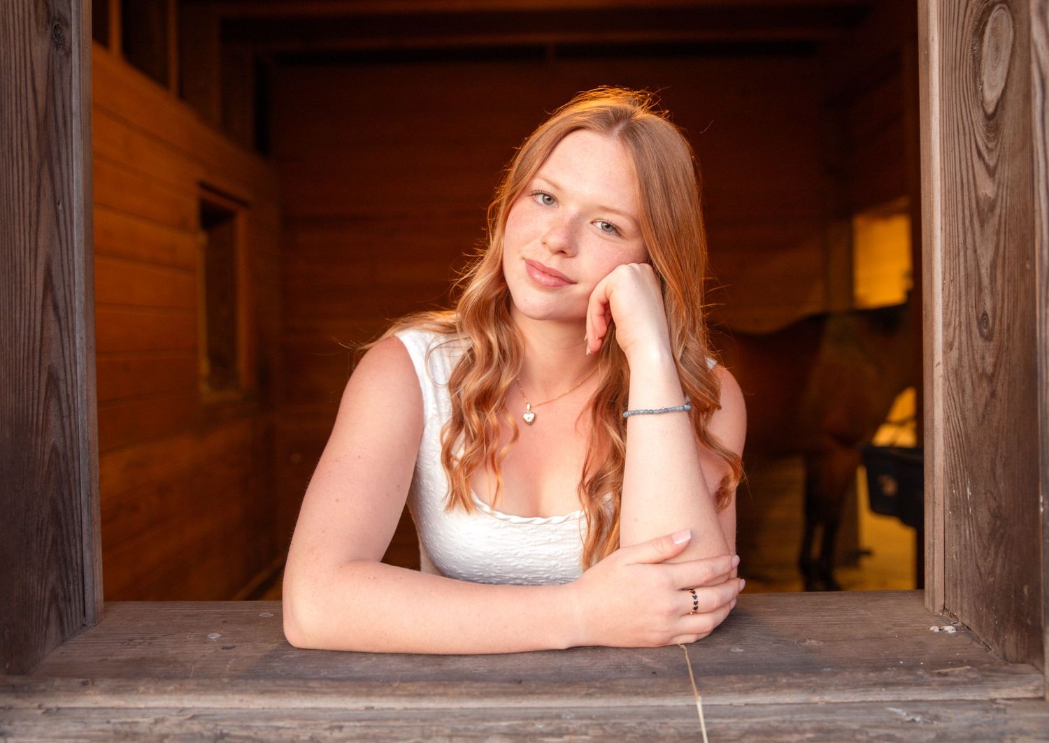 Young woman with red hair resting her chin on her hand, leaning on a wooden ledge in a warm barn setting.