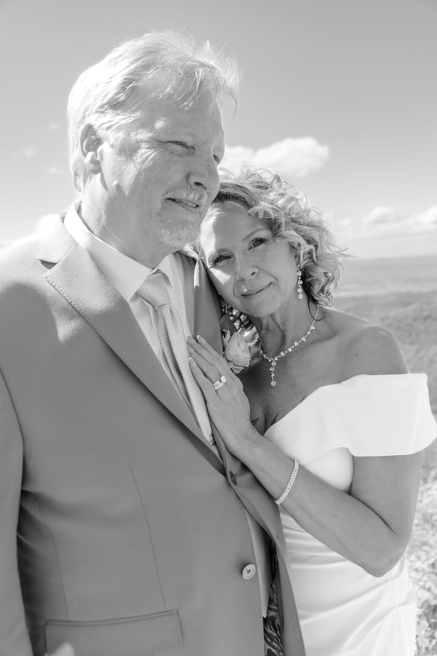 Elegant older couple in wedding attire pose together in black and white portrait with scenic sky backdrop.