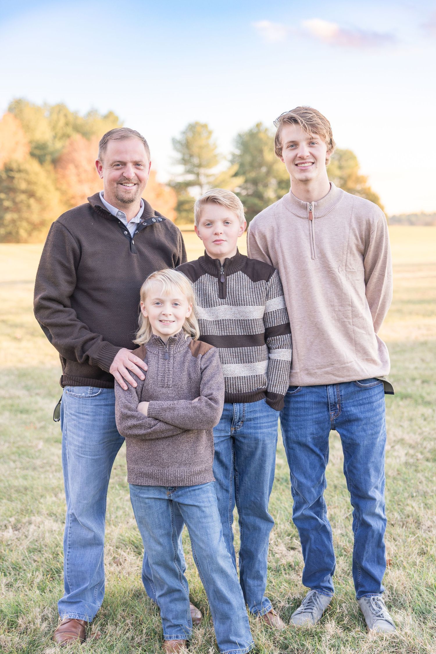 A family of four poses together in a grassy field during golden hour wearing casual sweaters and jeans.