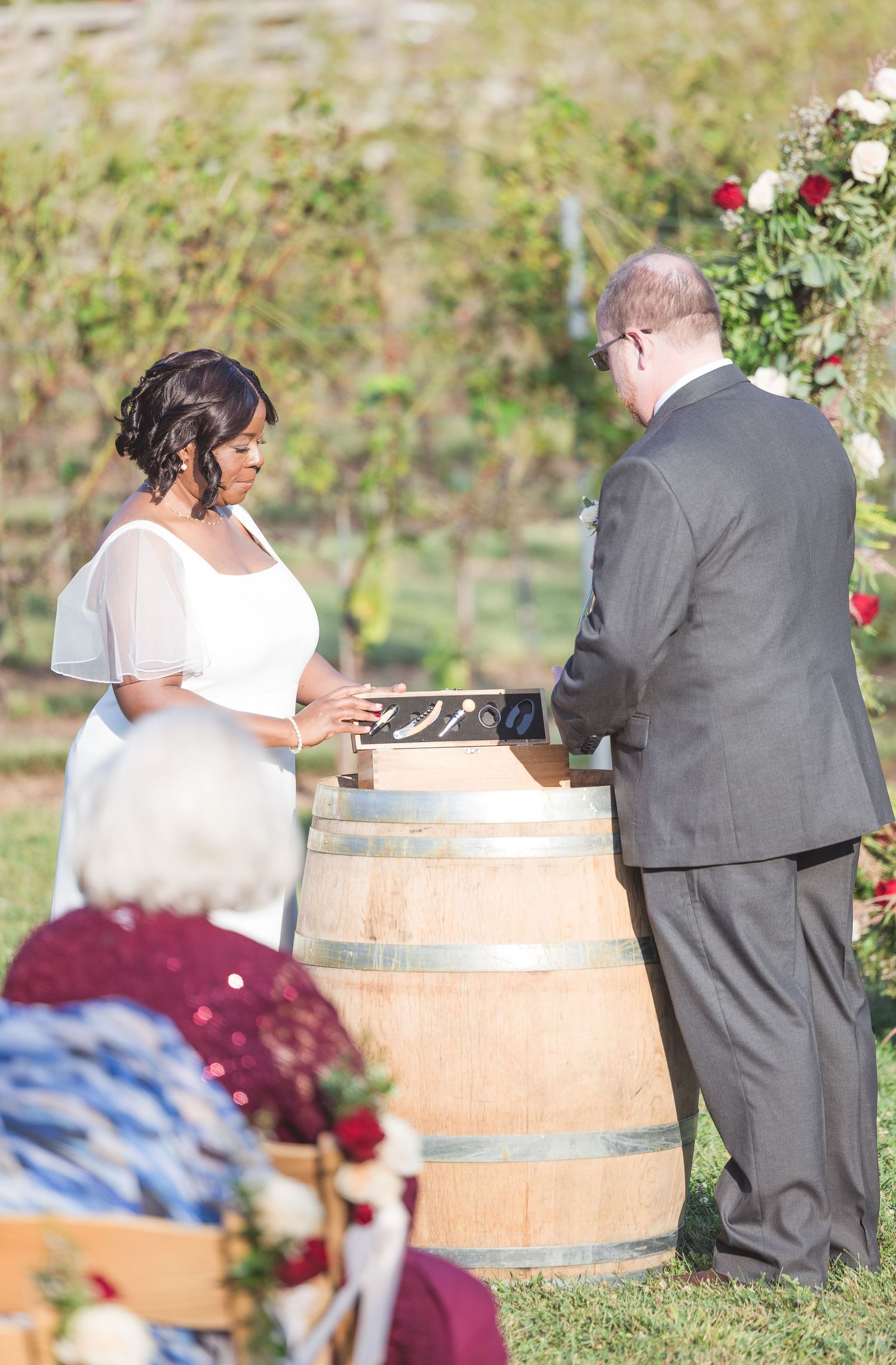 A wedding ceremony takes place in a vineyard setting with wooden barrel accents.