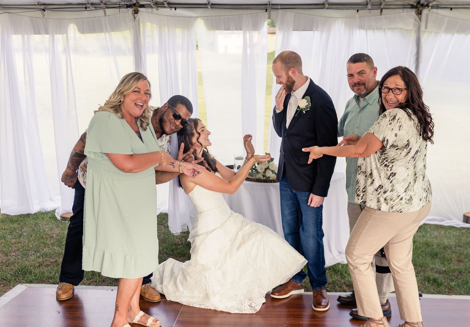 Bride in white gown dances joyfully with wedding guests under a white tent at an outdoor reception.
