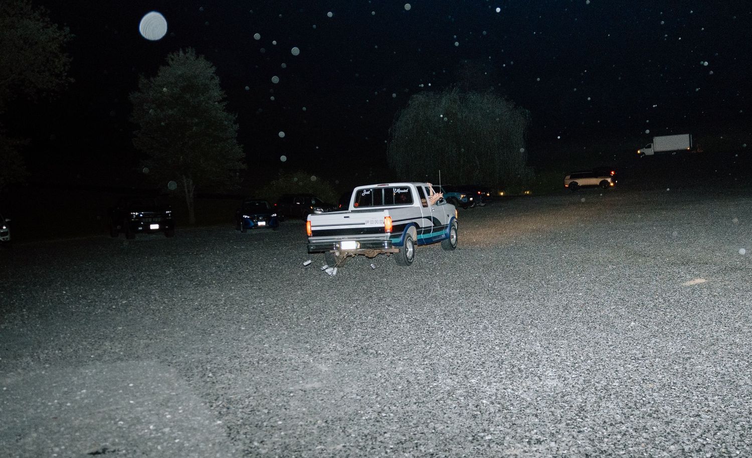A white pickup truck parked on a gravel lot at night under a full moon with trees in the background.