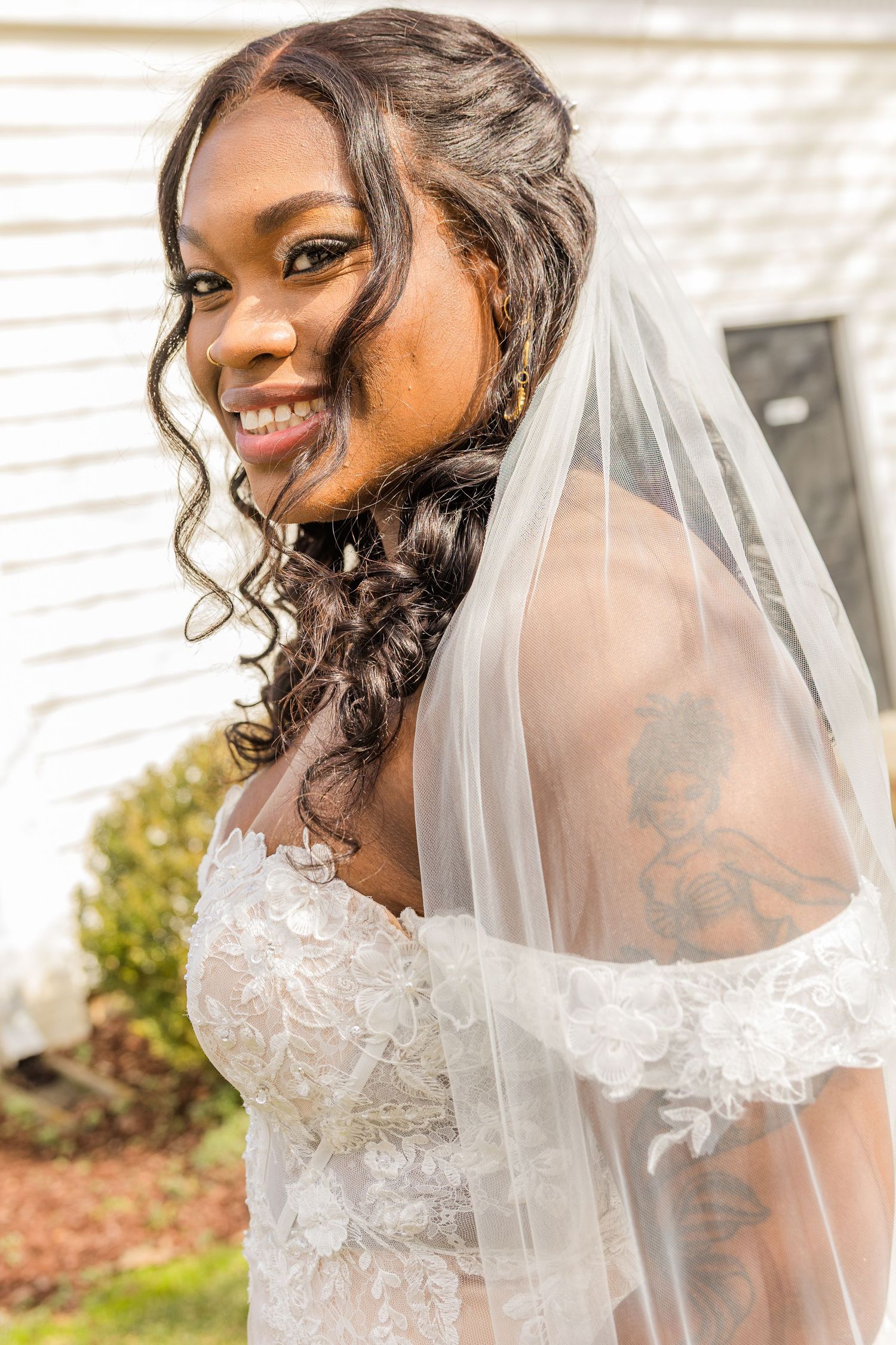 Beautiful bride wearing a white lace wedding dress and veil poses outdoors in natural lighting.
