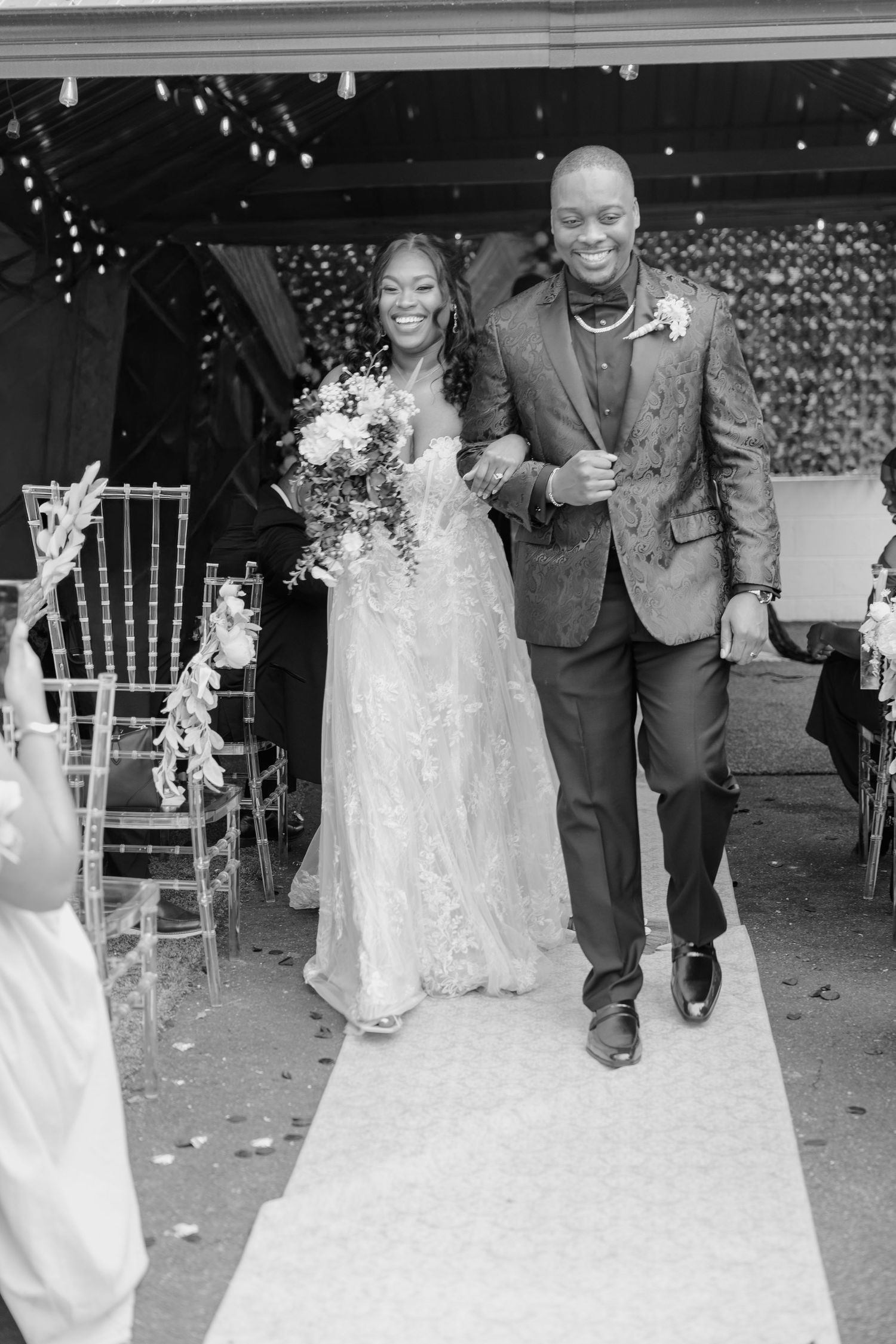 A joyful couple walks down a white aisle runner at their wedding ceremony, with the bride in a lace gown holding flowers.