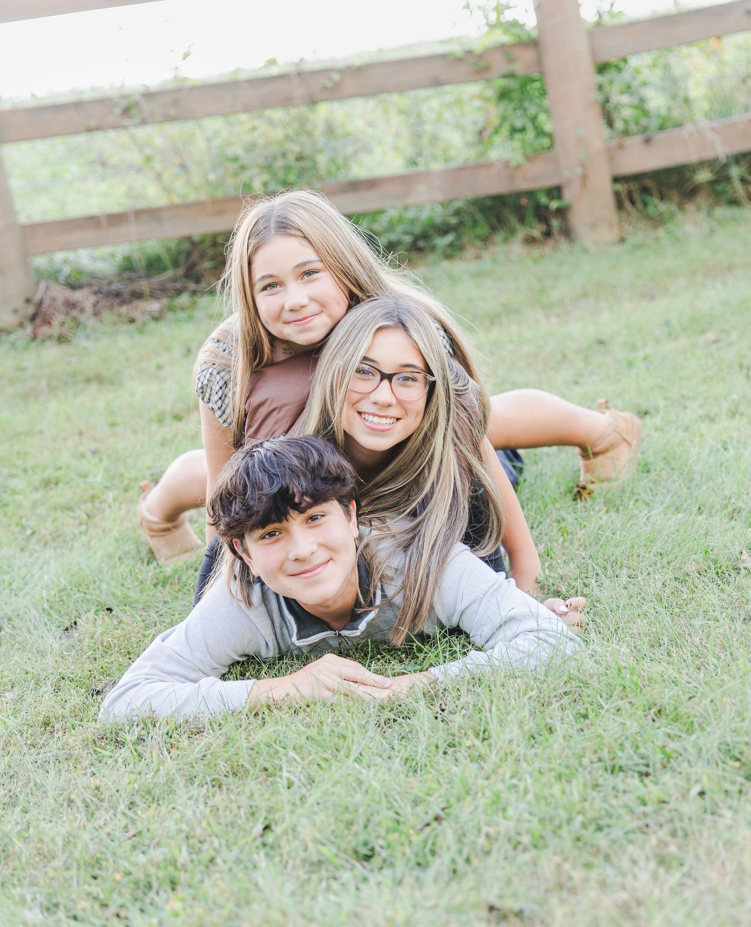 Three friends laying in green grass while posing playfully near a wooden fence on a sunny day.