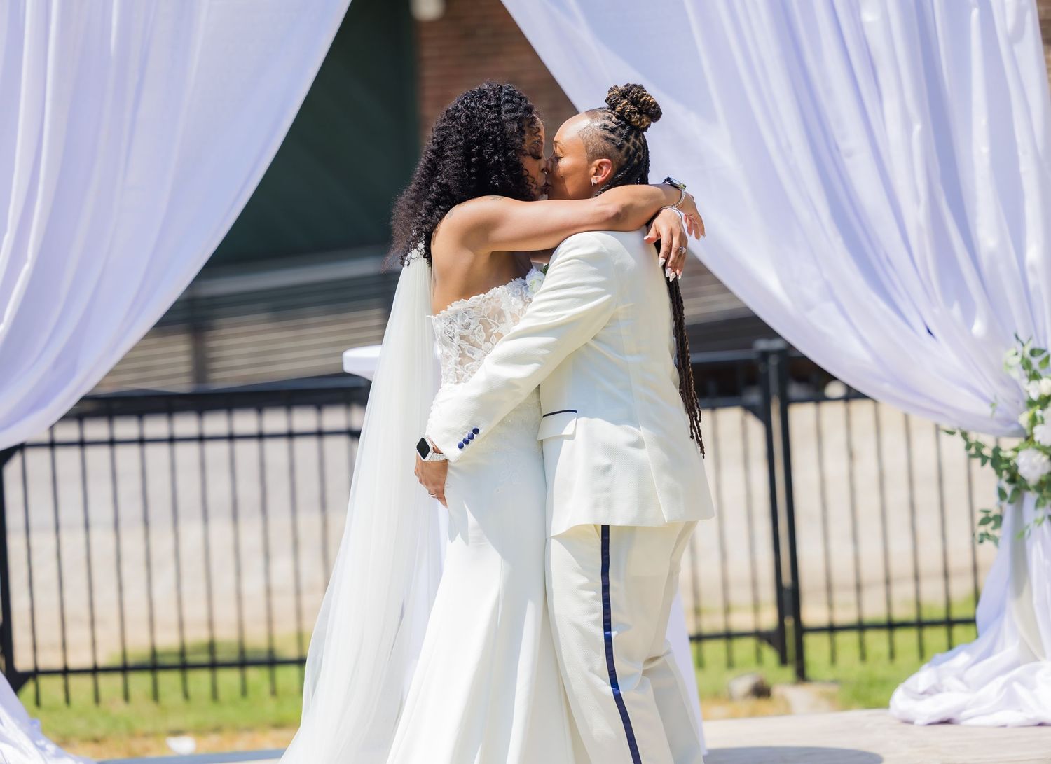 Newlyweds share first kiss as married couple under white draped wedding arch.