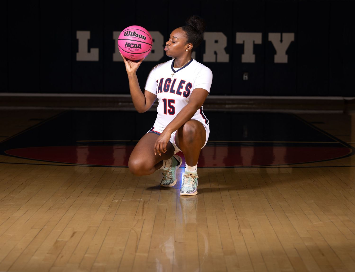 Basketball player in Eagles uniform poses with pink ball on wooden court.