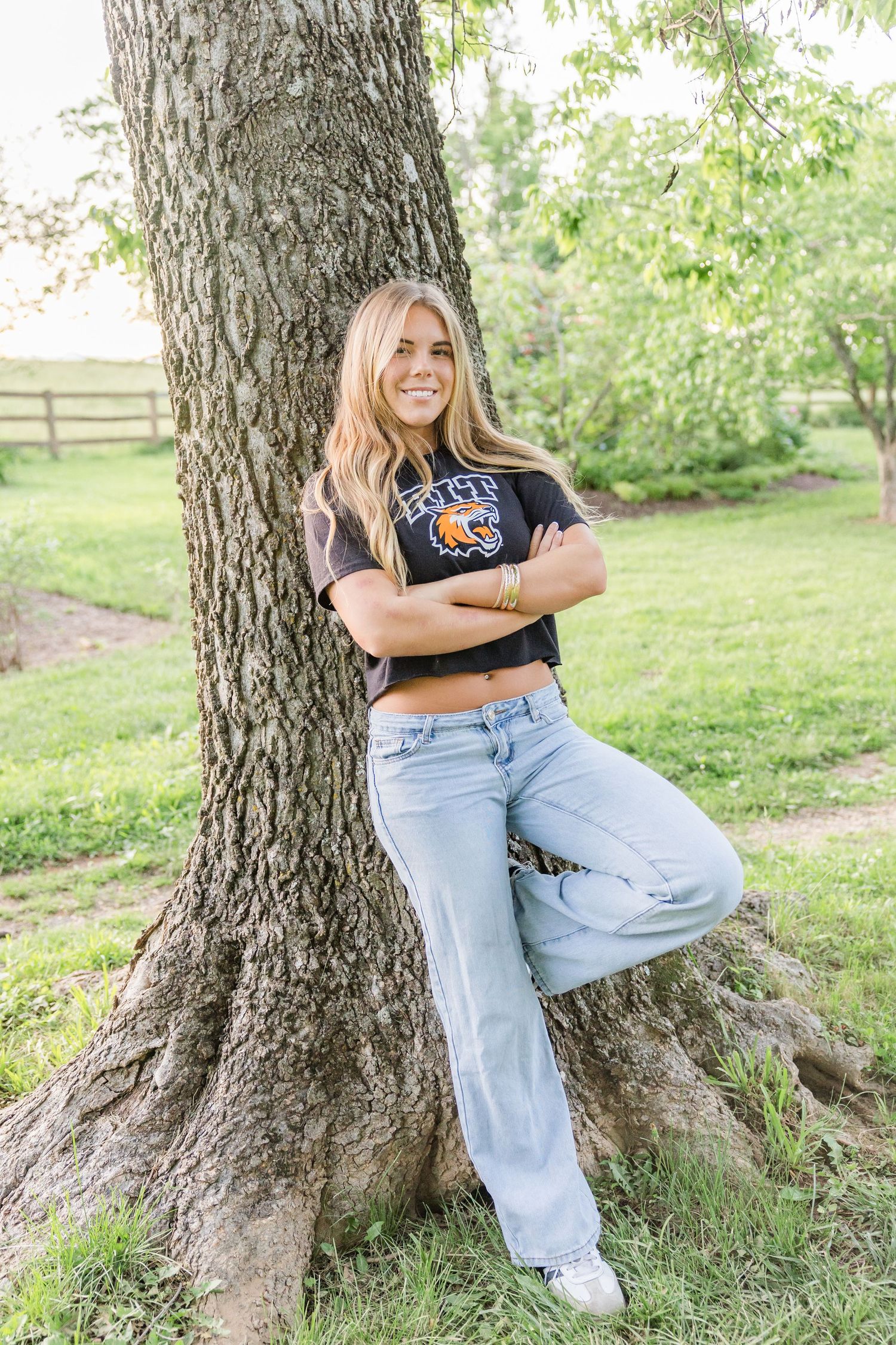 Woman in casual attire sitting and posing against a large tree trunk in a grassy field.