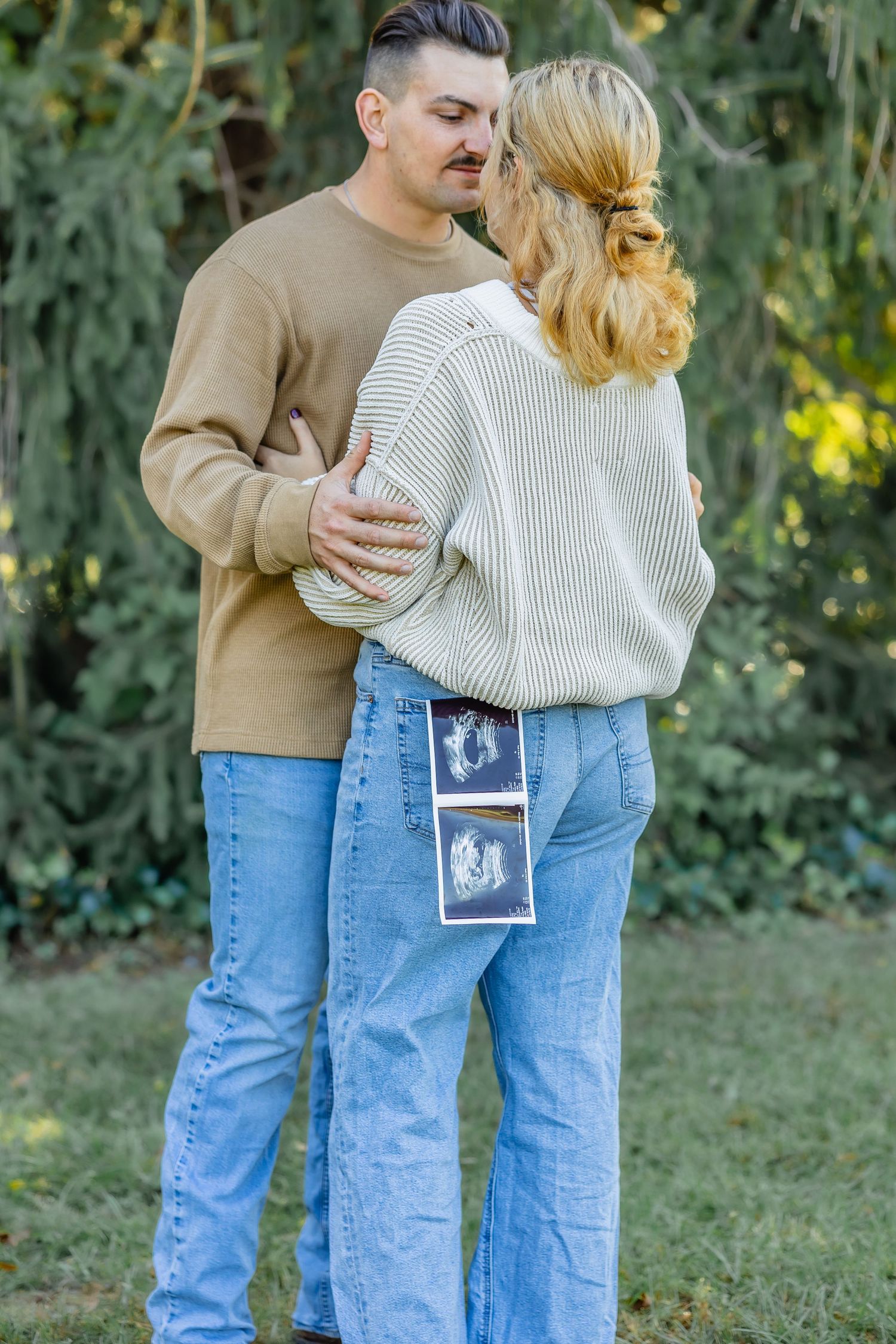Couple embracing outdoors with ultrasound photos visible in back pocket.