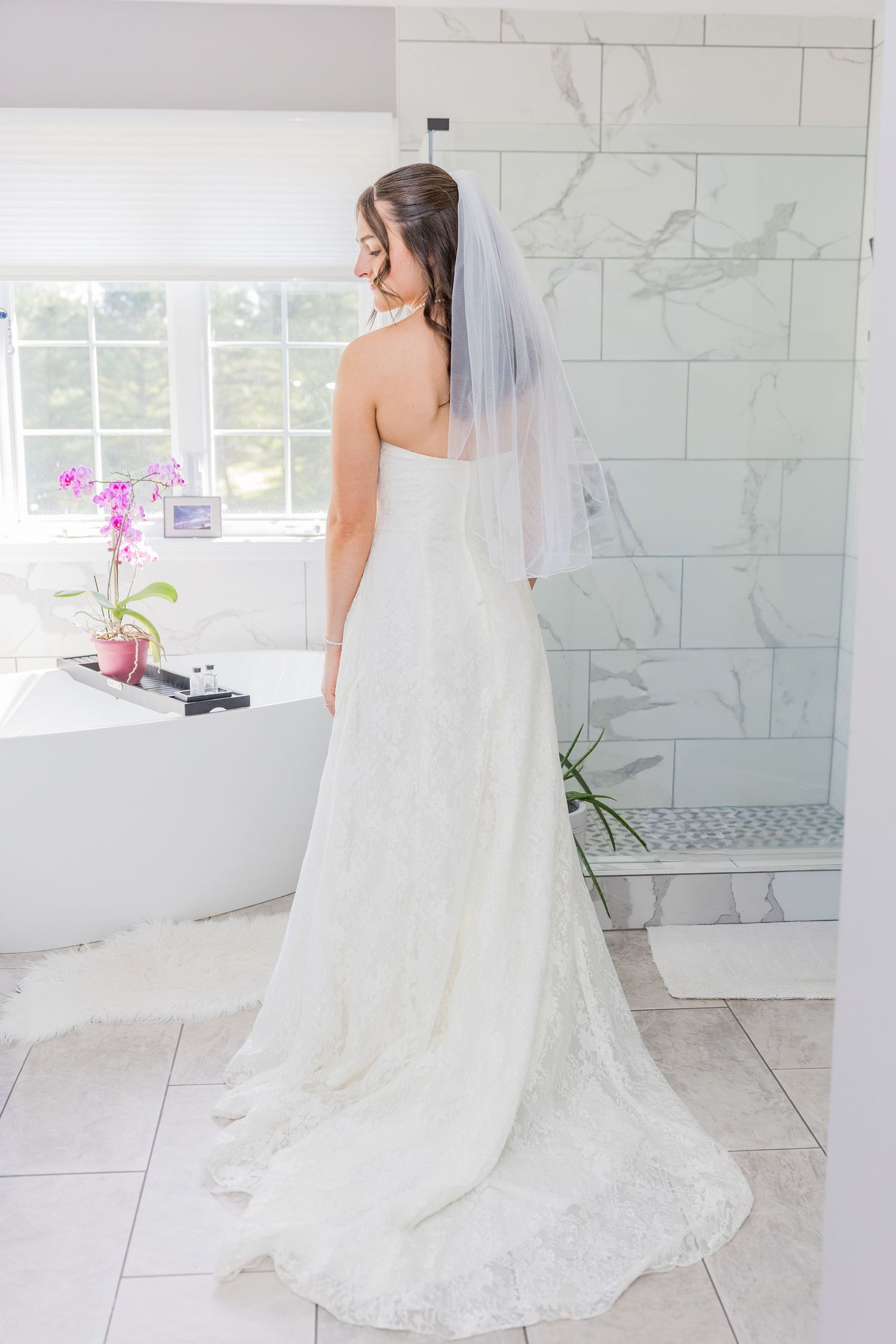 Bride in white lace wedding gown with veil stands in elegant marble bathroom near freestanding tub and orchid.
