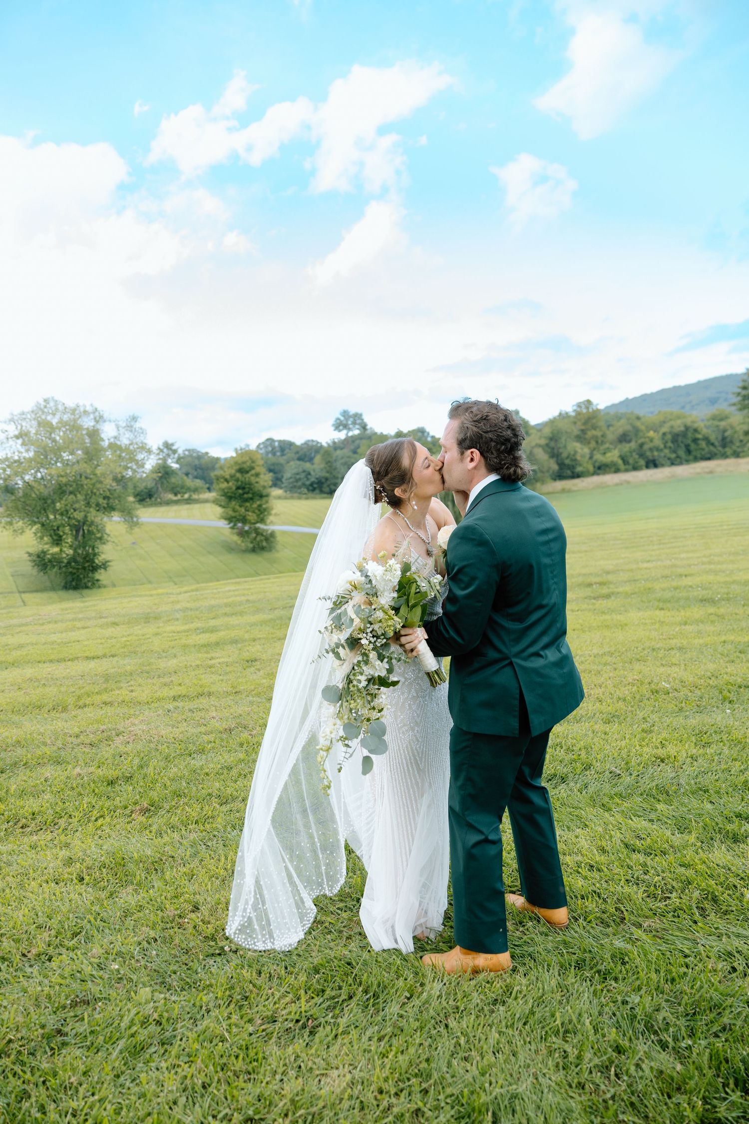 A bride and groom share a kiss on a lush green lawn, she holds a white bouquet, he wears a dark green suit.