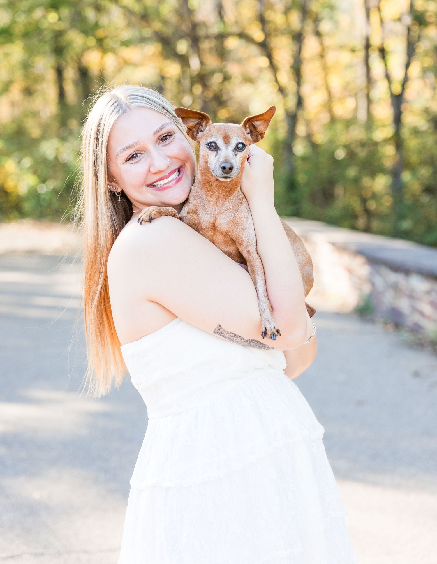 A person in white dress holds a small brown dog while standing outdoors.