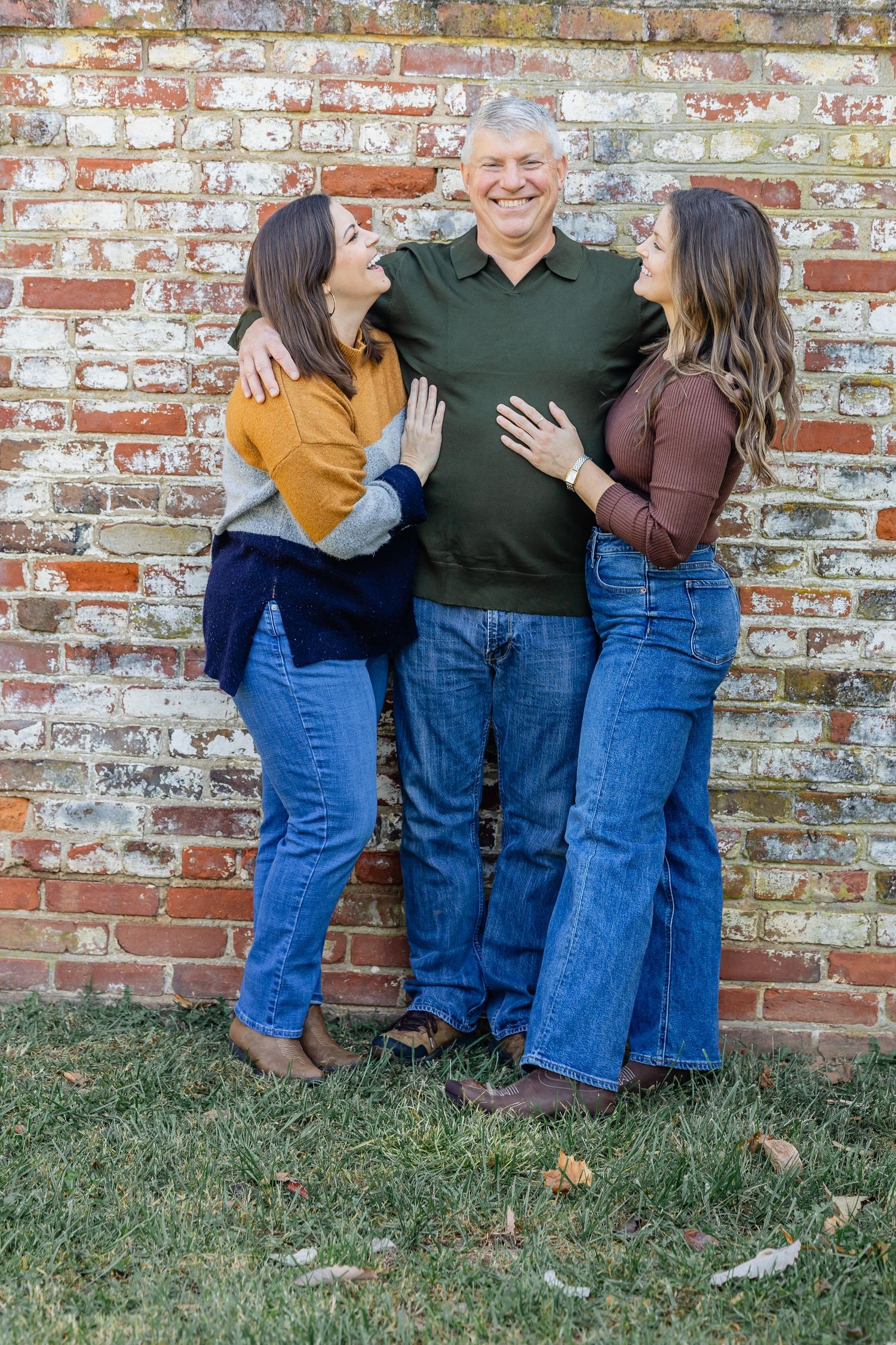 Three people pose casually together against a rustic brick wall background.