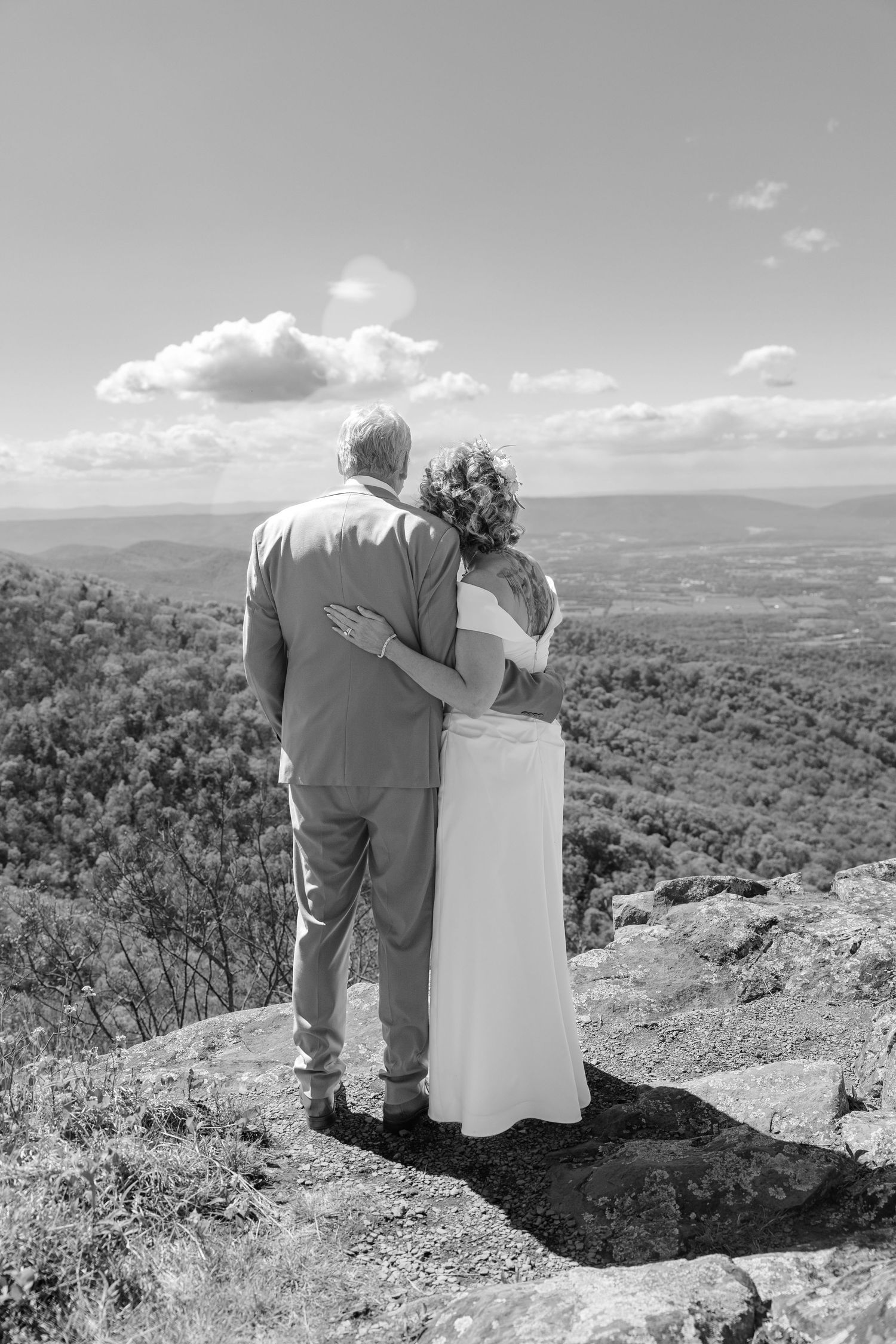 Black and white photo of a bride and groom embracing on a rocky overlook with a scenic mountain valley view.