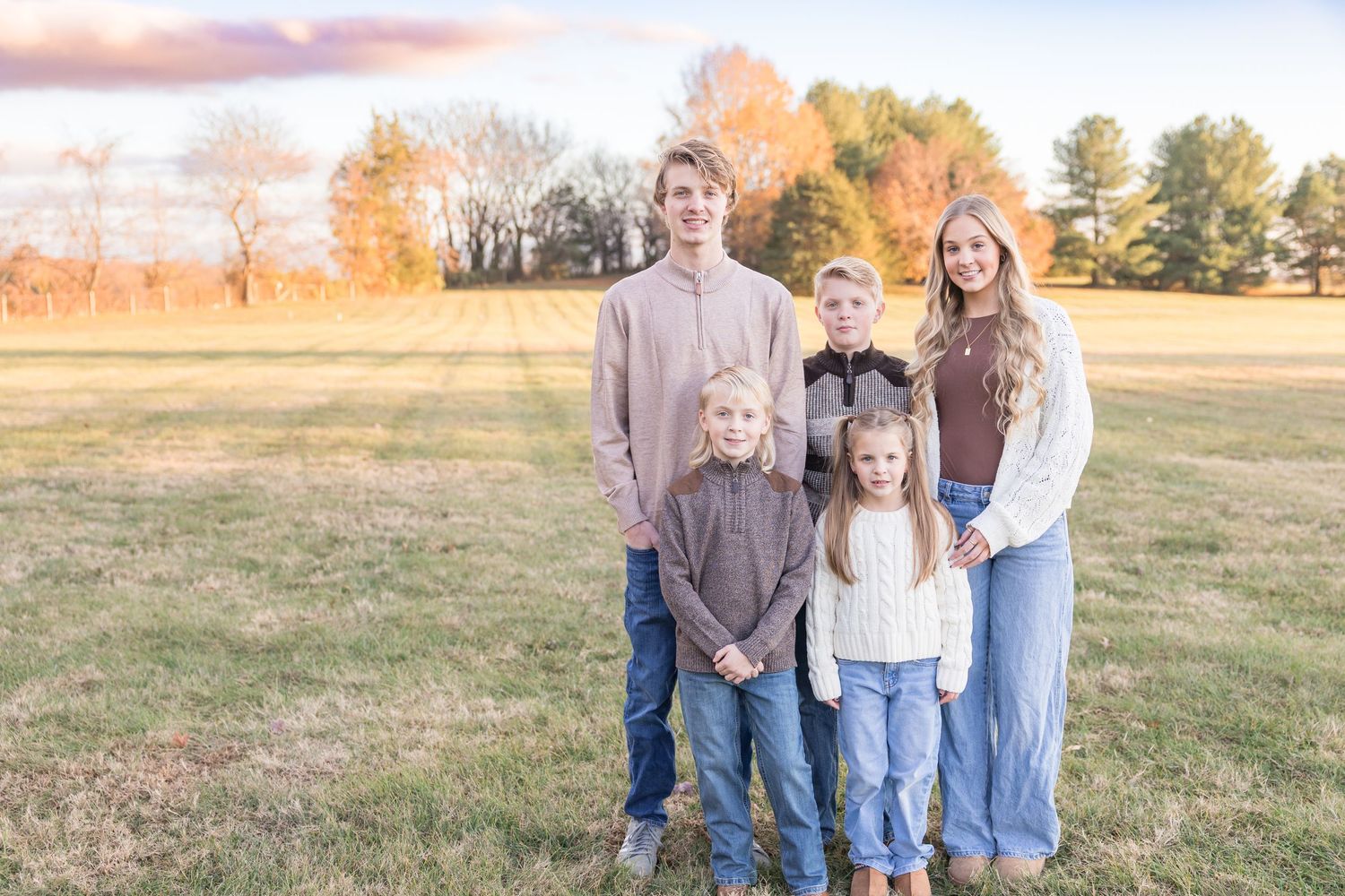 A family wearing coordinated neutral outfits poses together in a golden field at sunset.