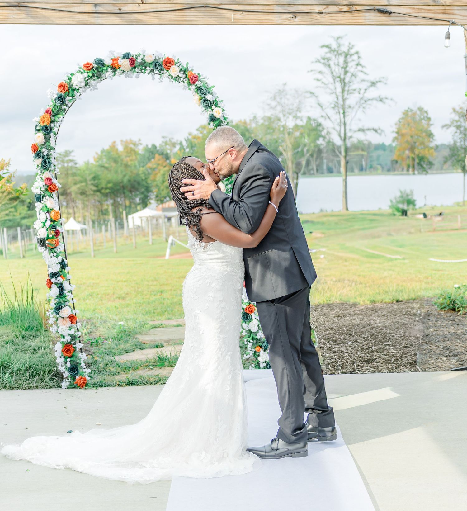 A romantic wedding kiss under a floral arch with a lake and trees in the background.