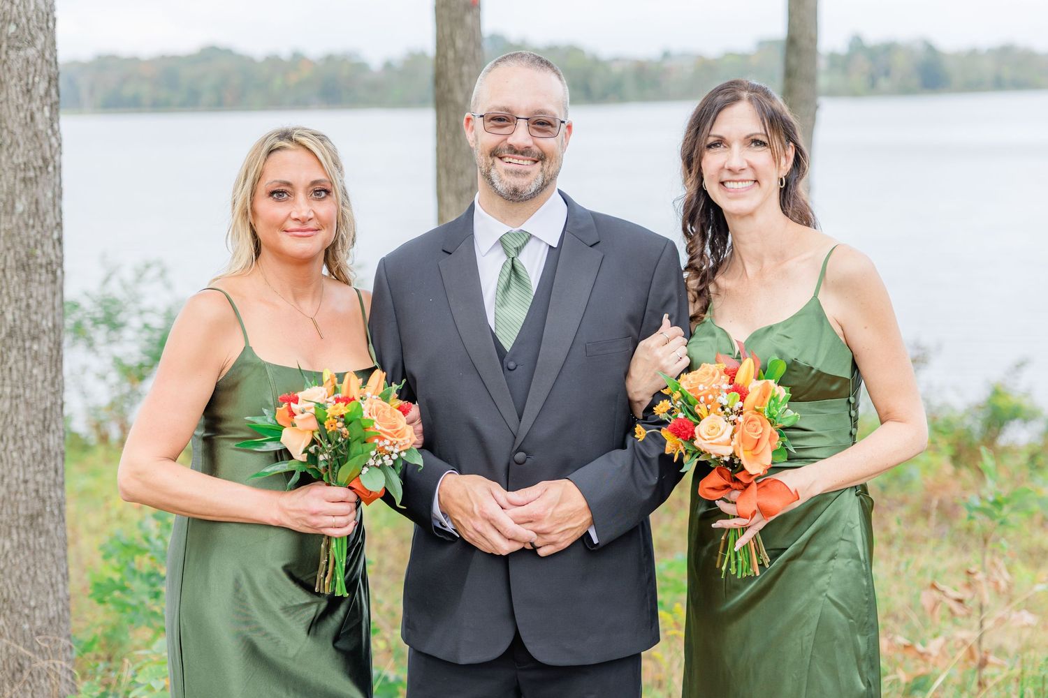 Wedding party in green dresses and gray suits poses outdoors near water.