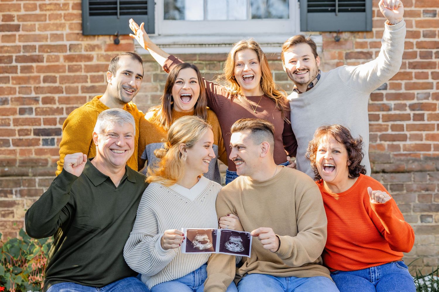 A joyful family group shows ultrasound photos while celebrating together on outdoor steps.