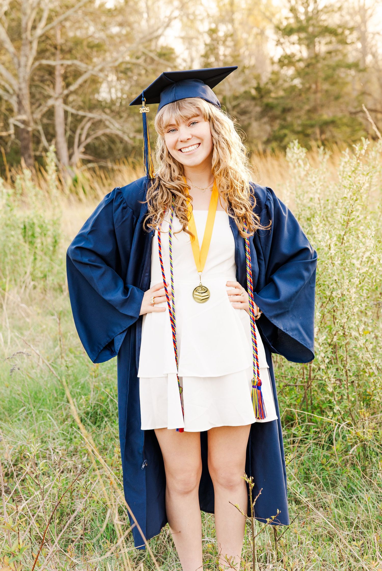 Graduate in blue cap and gown poses joyfully in sunny field.
