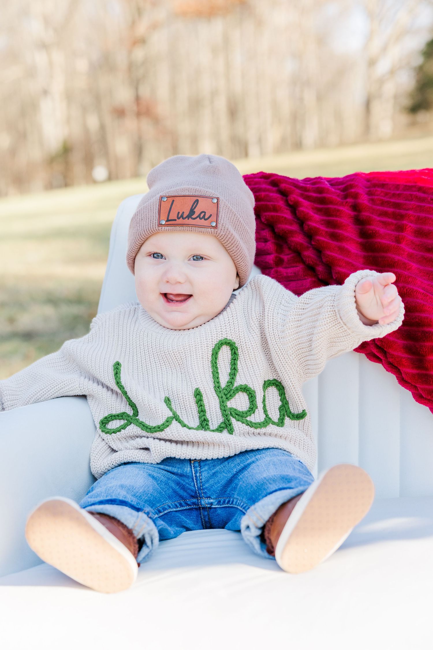 Smiling baby in winter outfit poses on white bench with red blanket.