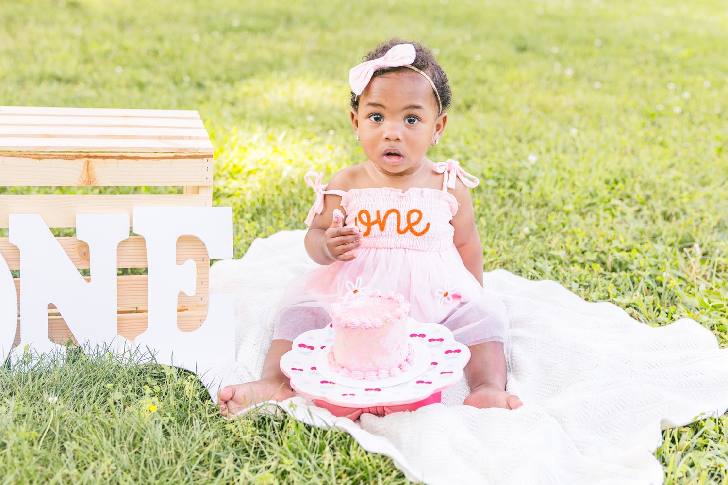 Baby in pink dress plays on white blanket during outdoor photoshoot.