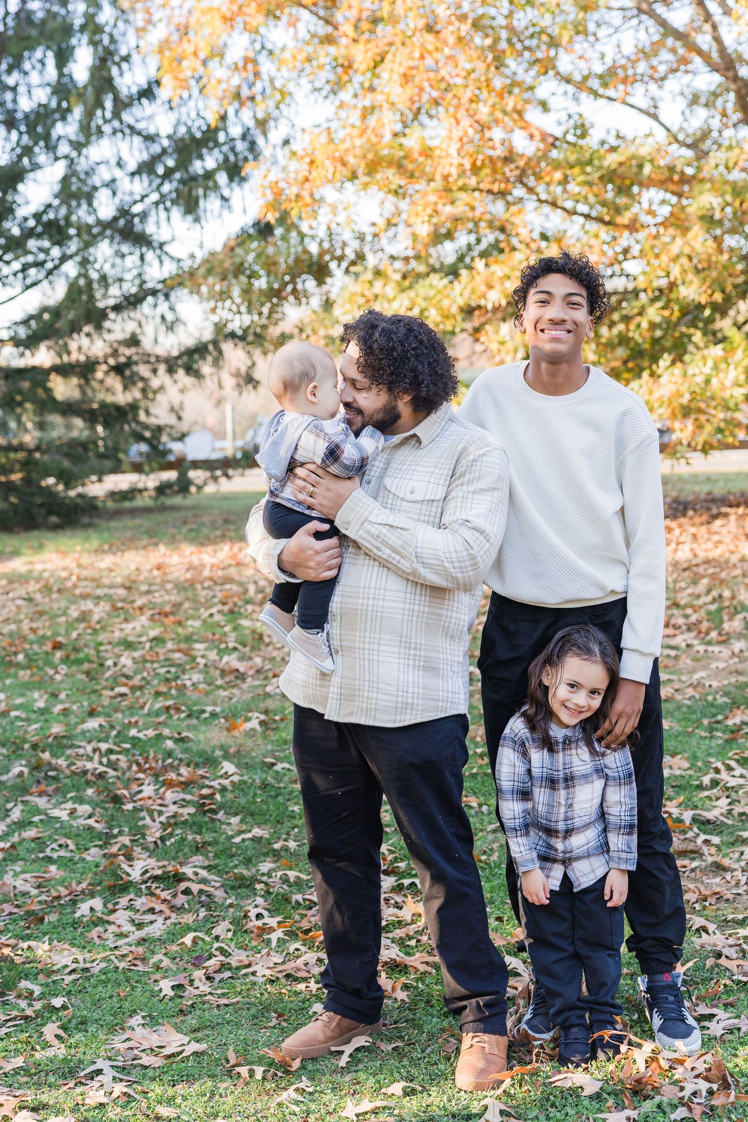 A family group poses together outdoors on an autumn day with colorful fall foliage in the background.
