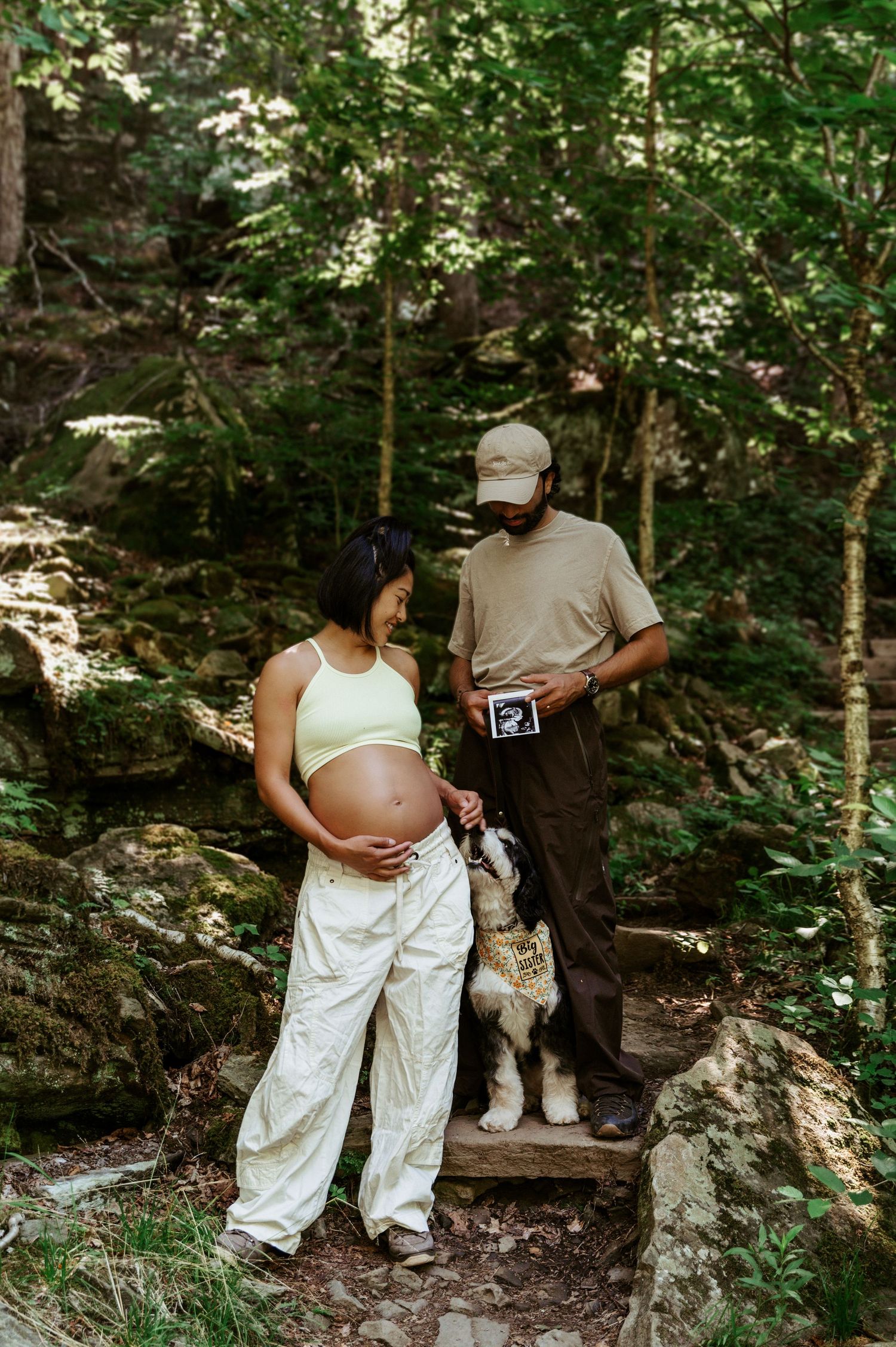 Pregnant woman and man with dog holding ultrasound photo on rocky forest trail for maternity announcement.