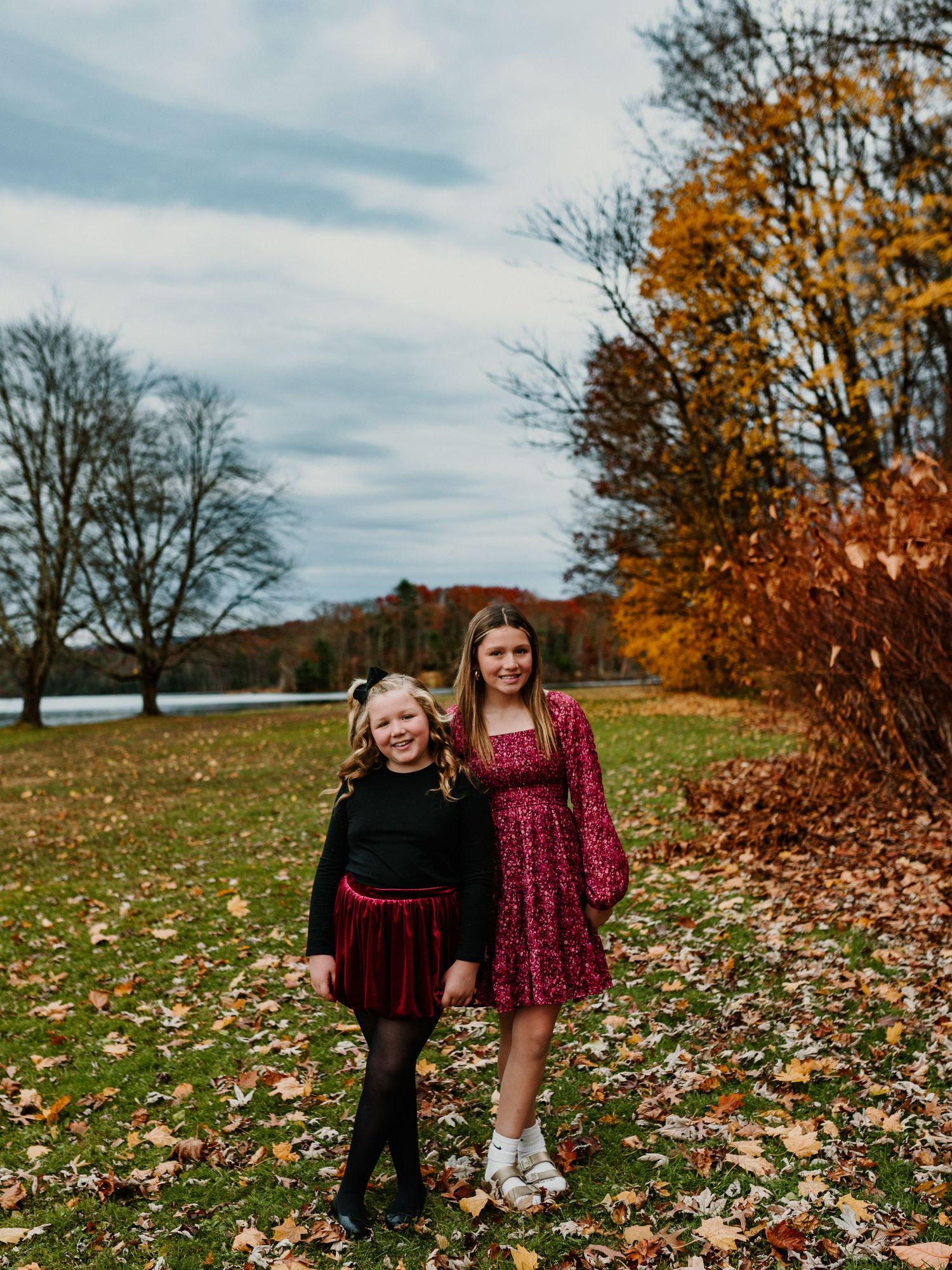 Two people in burgundy velvet dresses stand among fallen autumn leaves on a grassy hillside during fall.