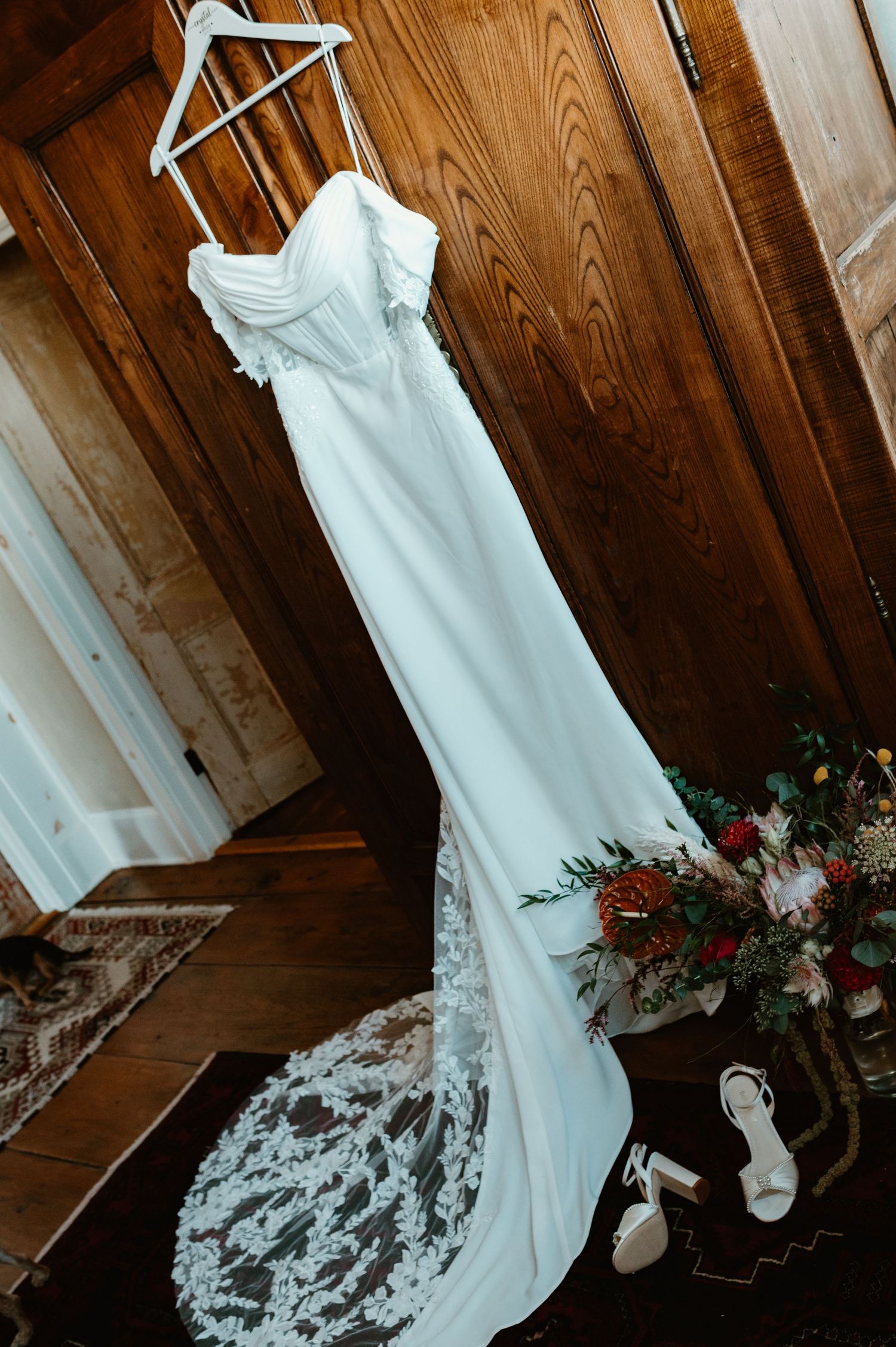 A wedding dress hangs against a rustic wooden wall with white lace details and floral arrangements.