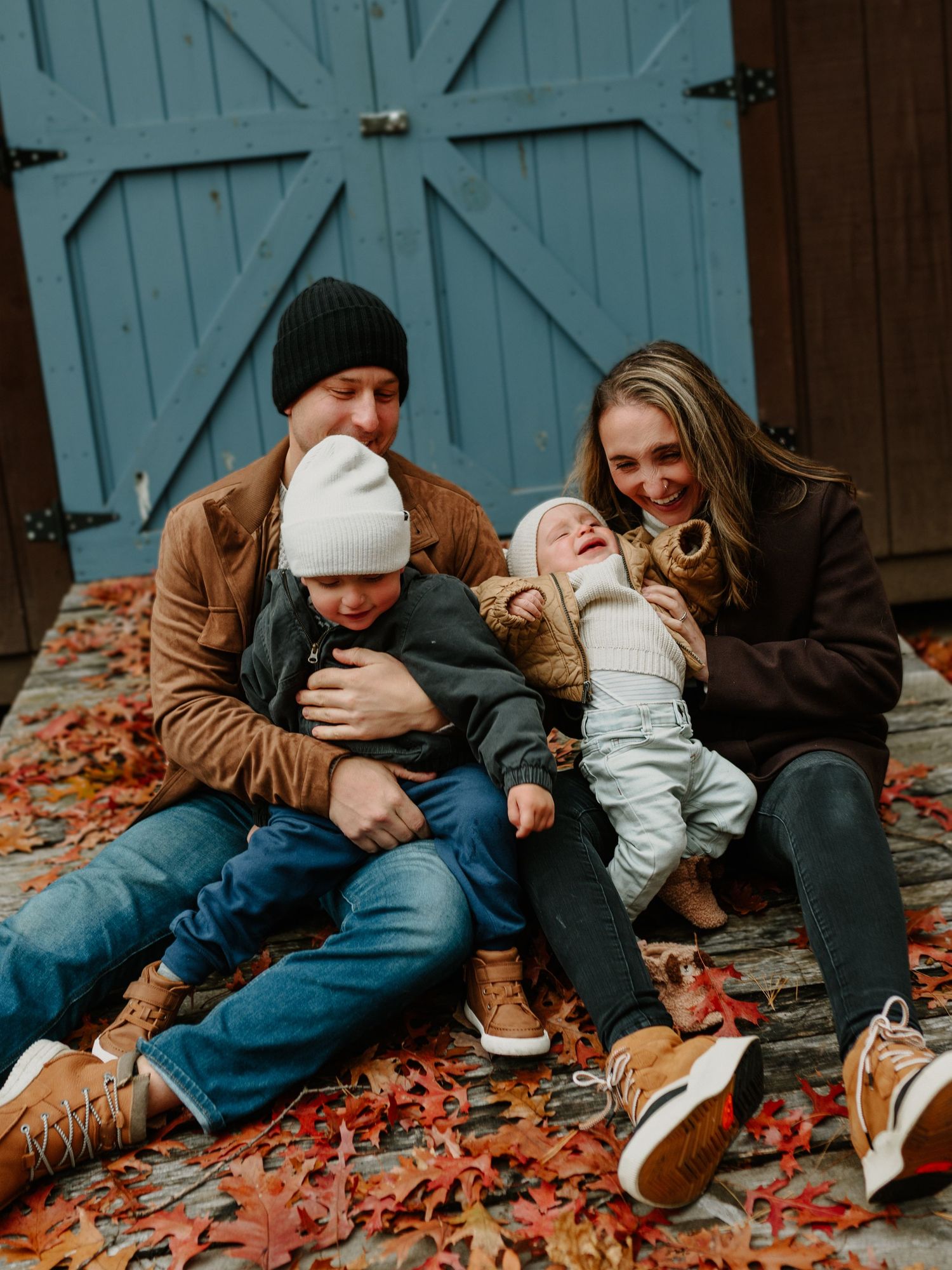 A family sits together on fallen autumn leaves in front of a turquoise barn door wearing cozy winter clothing.