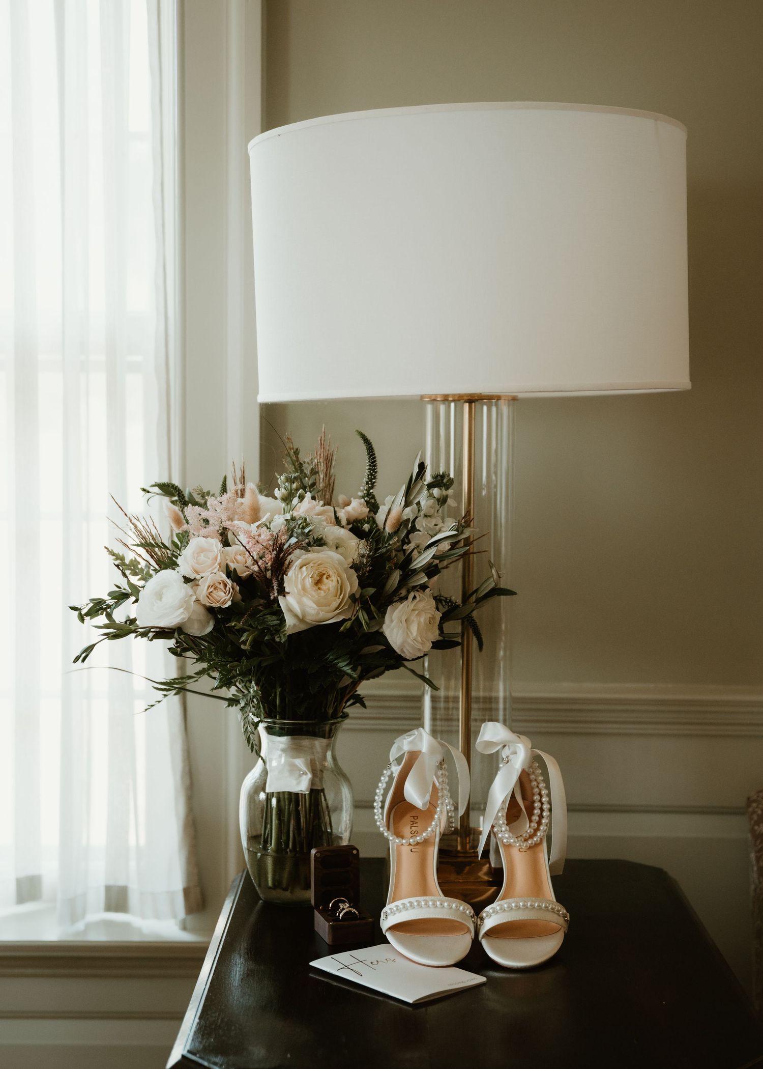 Elegant white bridal shoes and floral arrangements displayed on windowsills with soft natural lighting.