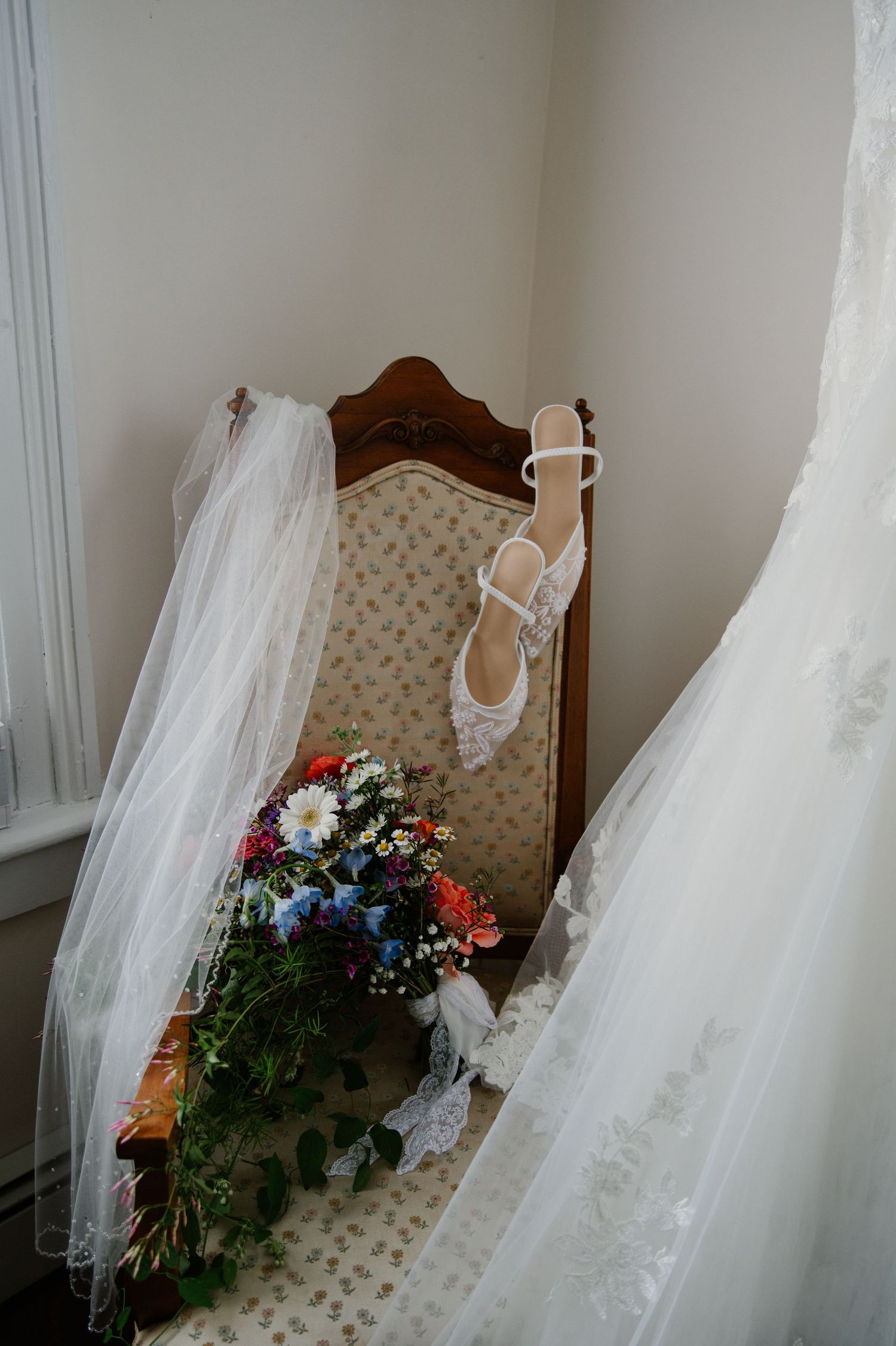 Bridal flat lay with lace heels on vintage chair, colorful wildflower bouquet, flowing white gown and veil in soft natural light.