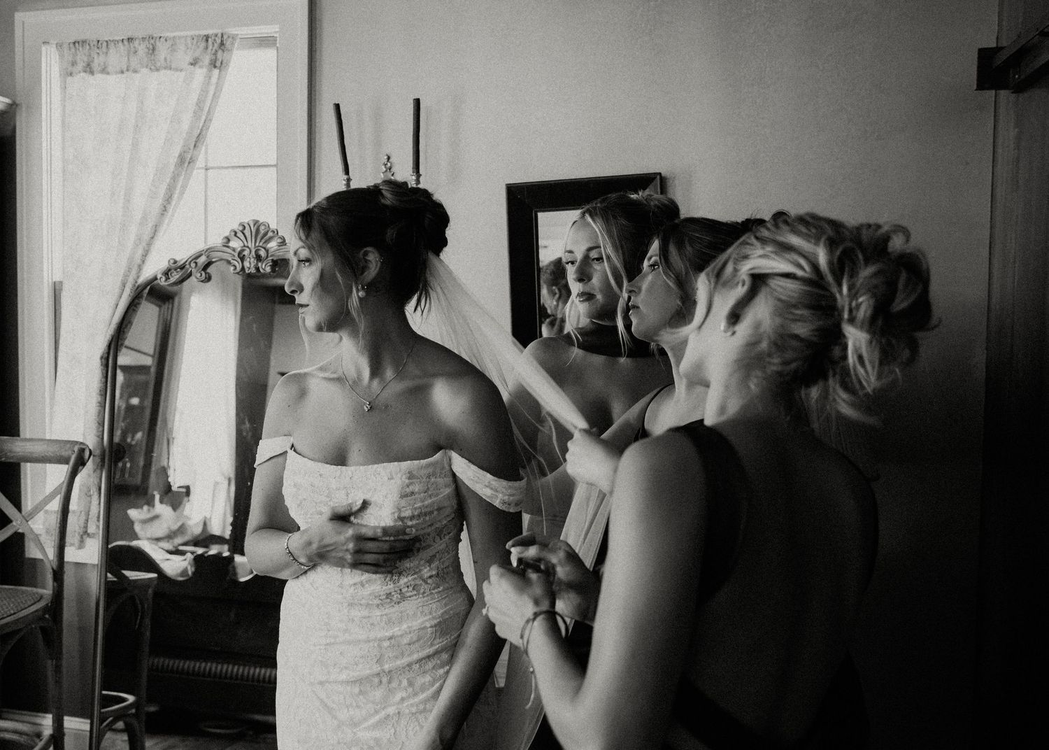 Black and white photo of a bride in a strapless gown being helped by a bridesmaid, with a mirror reflection visible.