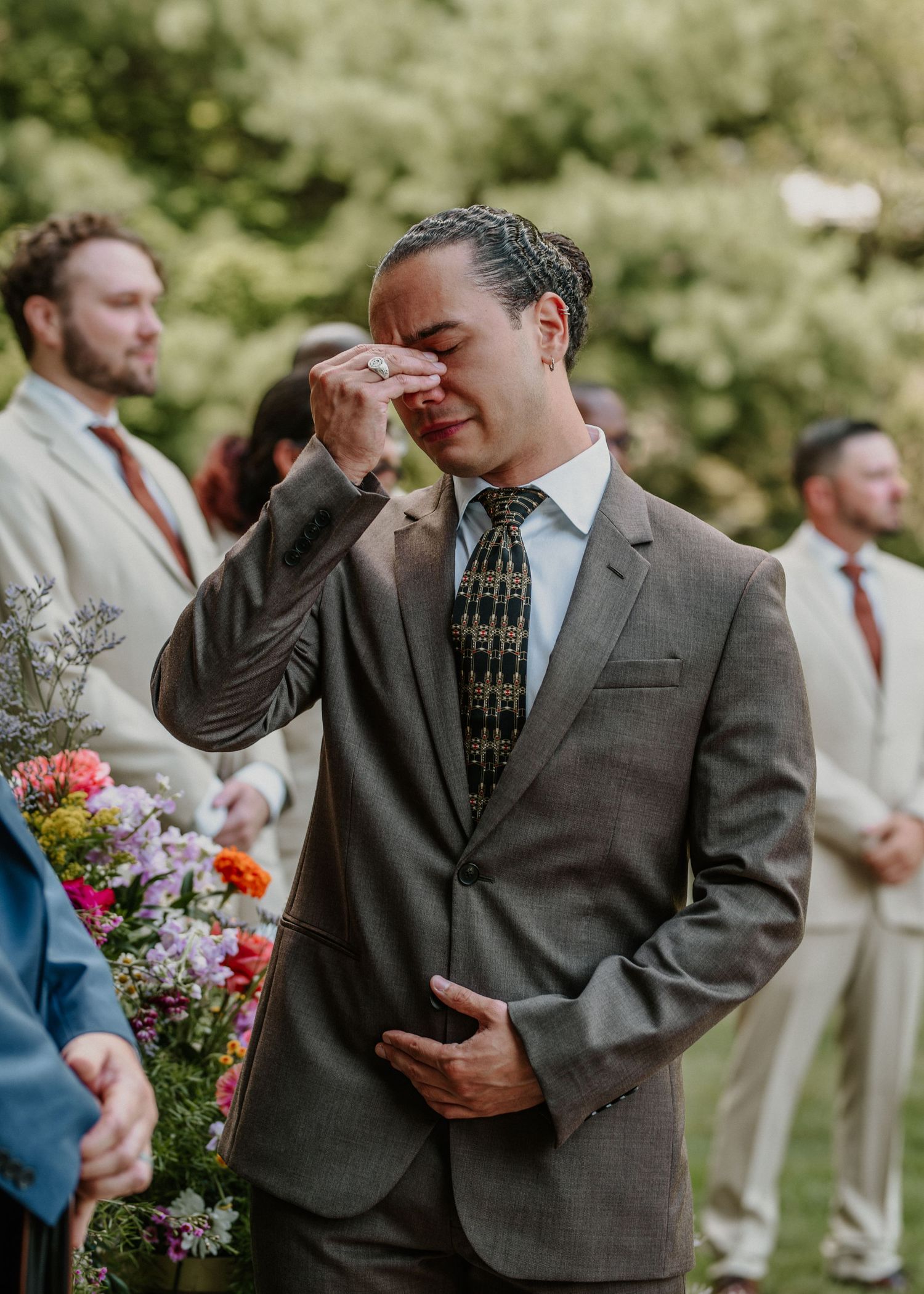 Emotional groom wiping tears at outdoor wedding ceremony wearing gray suit and patterned tie with groomsmen in background.