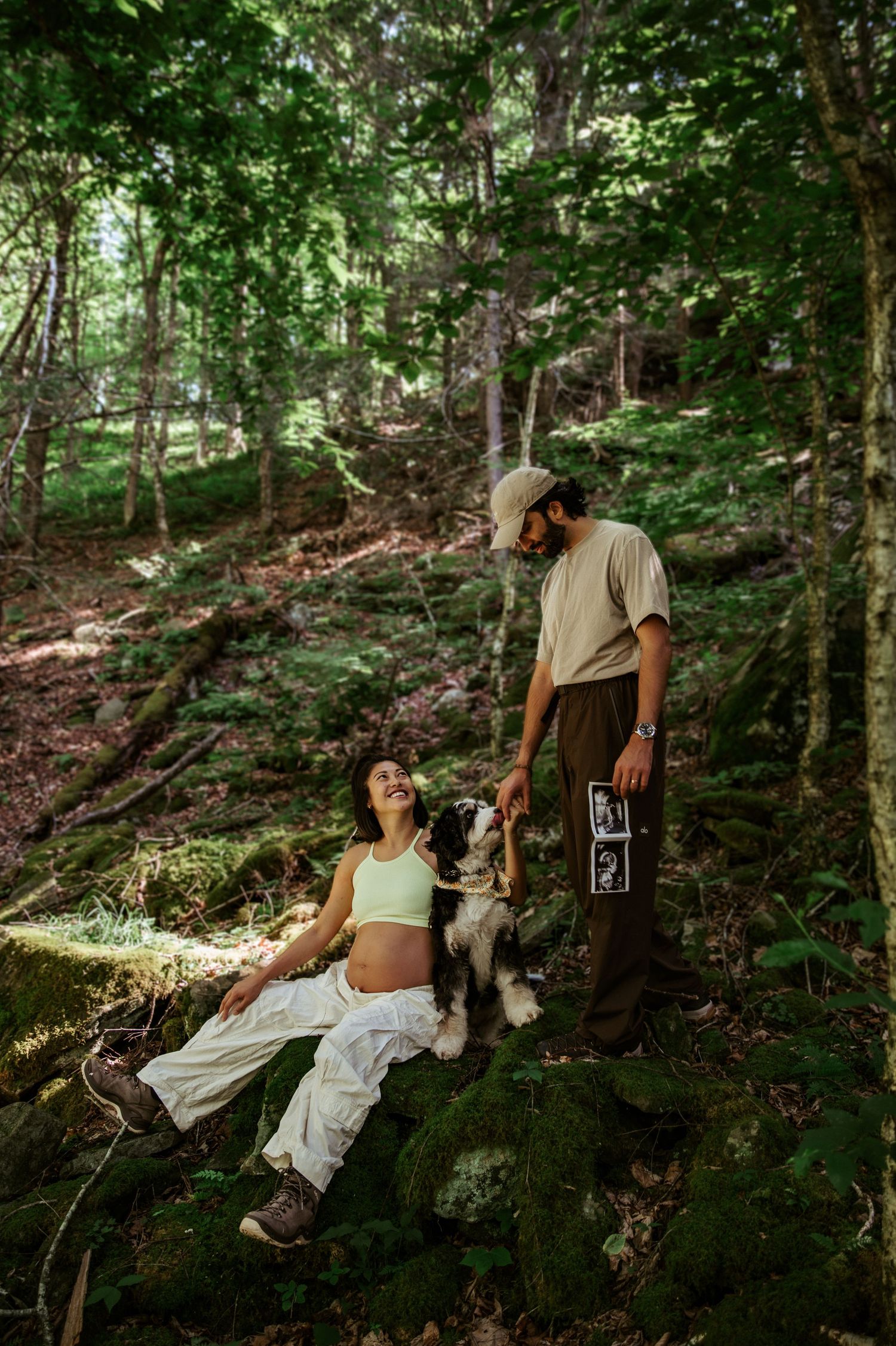 Pregnant woman sitting on mossy rocks in forest with partner holding ultrasound photo and Border Collie dog.