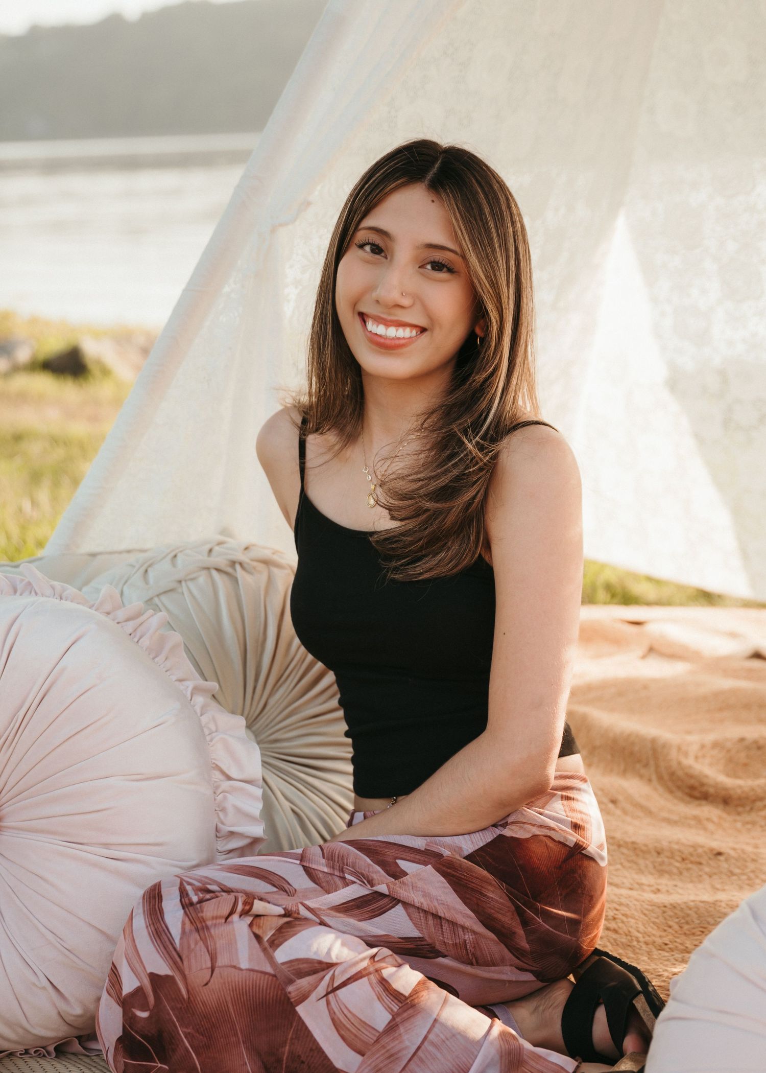 Young woman smiling outdoors on sandy beach, wearing black tank top and floral pants, seated near a white teepee tent.
