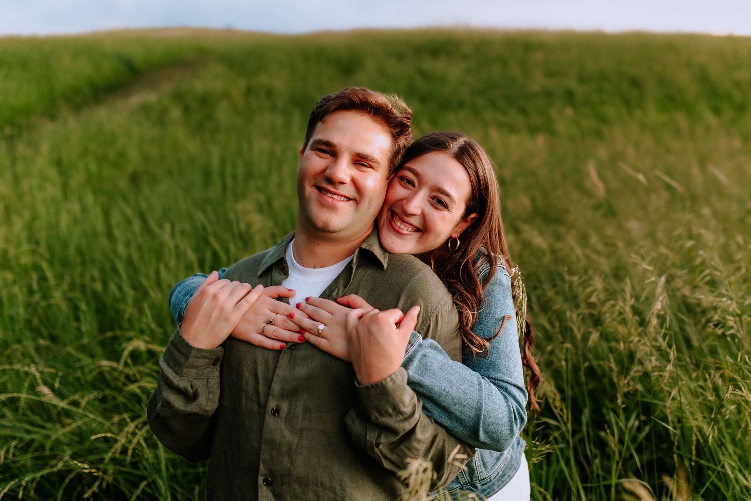 Happy couple embracing in a green field, woman hugging man from behind, both smiling outdoors on a sunny day.