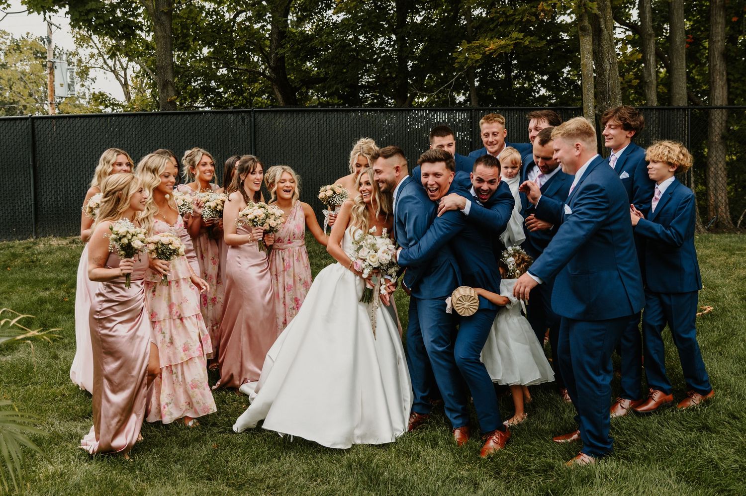 Wedding party members in pink dresses and navy suits pose playfully together outdoors for group photos.