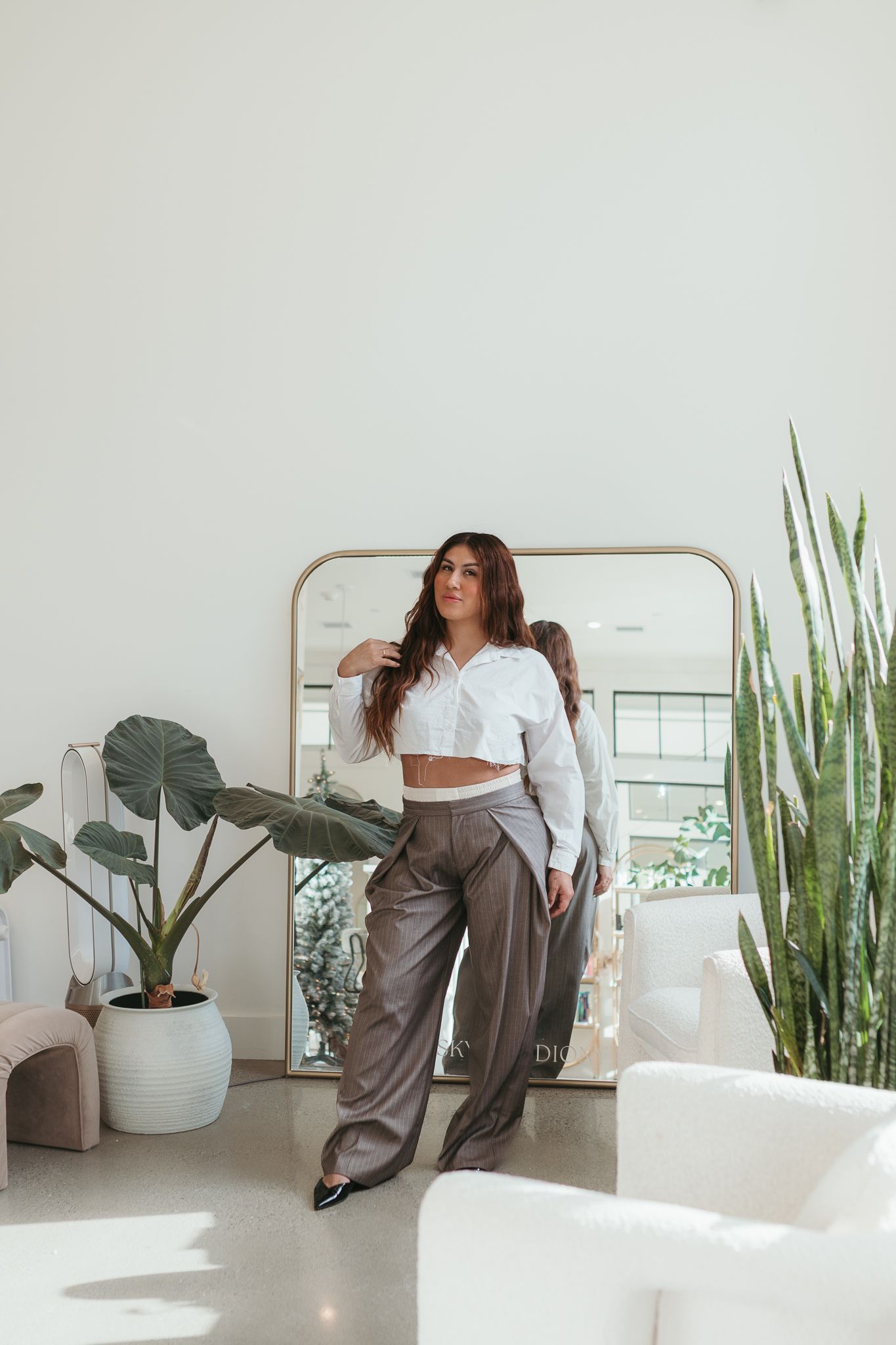 Minimalist white room interior with tall plants and someone lounging in casual grey attire near modern furniture.
