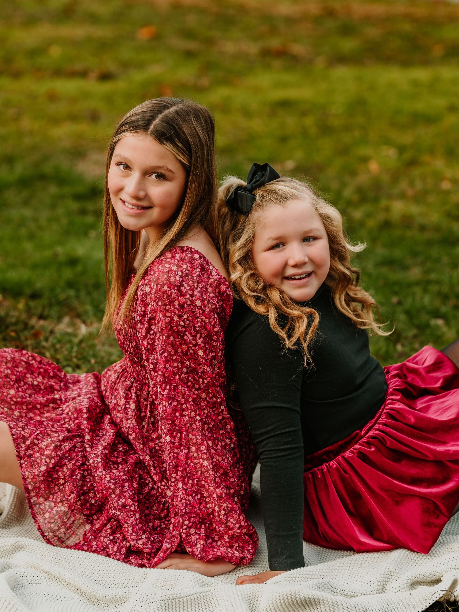 Two children in sparkly holiday dresses sit together on a white blanket outdoors on grass during an autumn photoshoot.