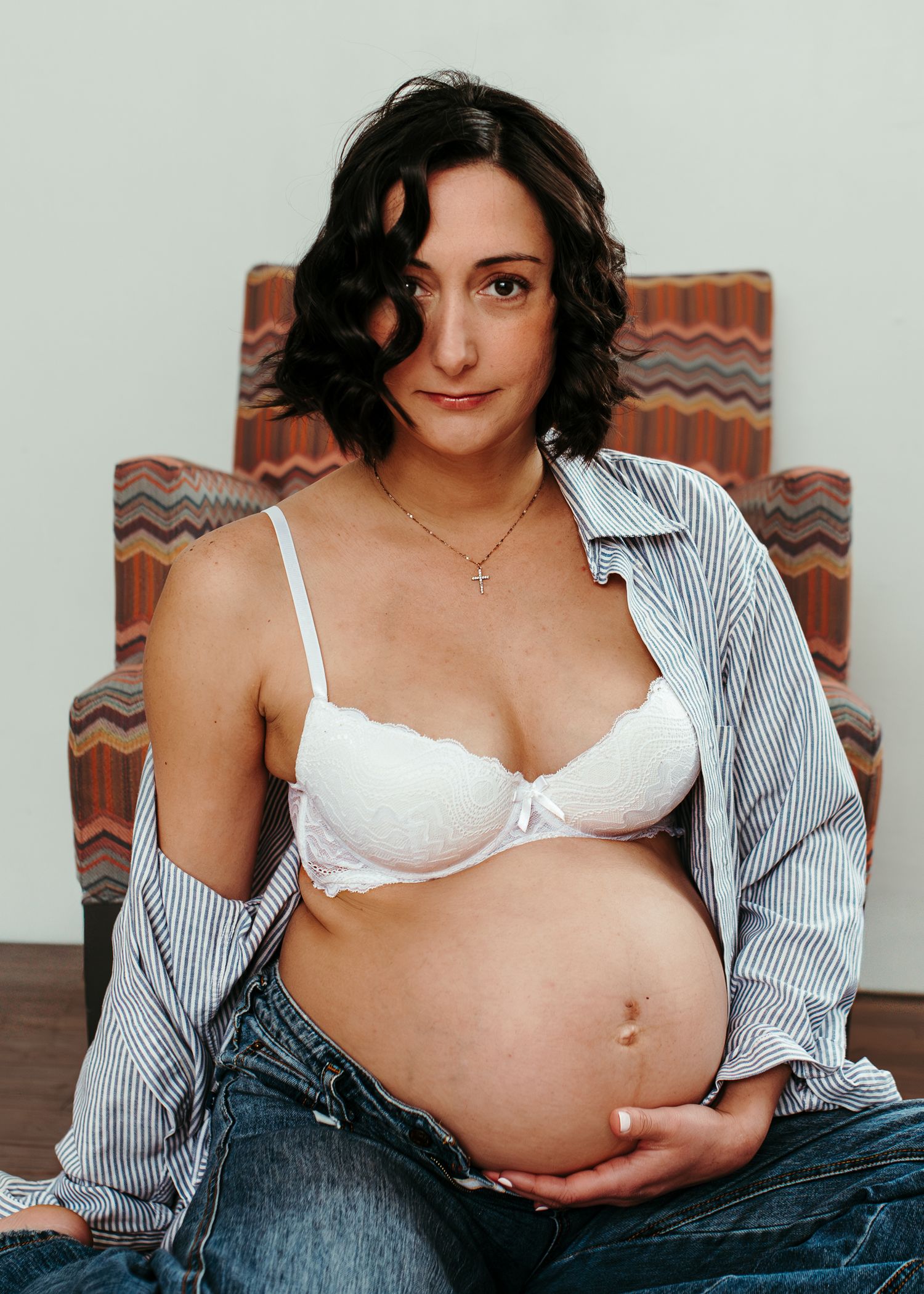 Pregnant woman in white bra and open striped shirt sits in colorful chair, cradling baby bump in maternity photo.