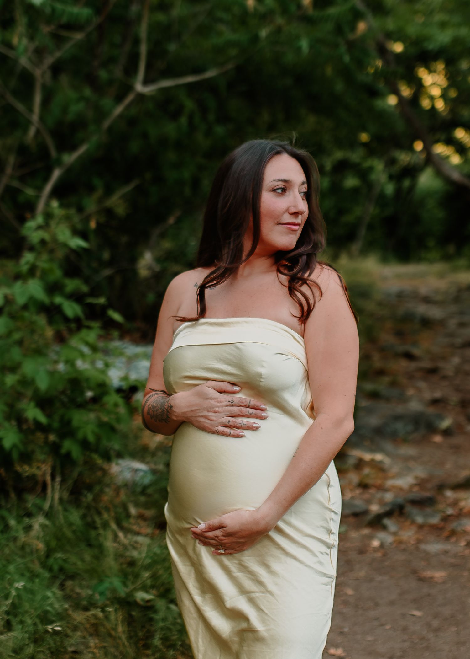 Pregnant woman in cream strapless dress cradling her baby bump outdoors on a forest path.