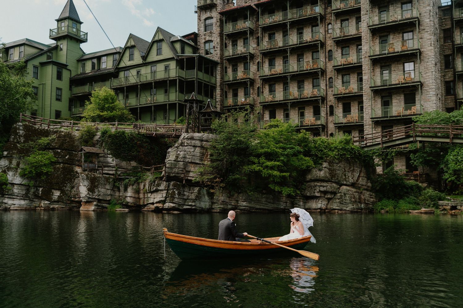 Bride and groom rowing a wooden boat on a lake beside a grand stone resort building at Mohonk Mountain House.