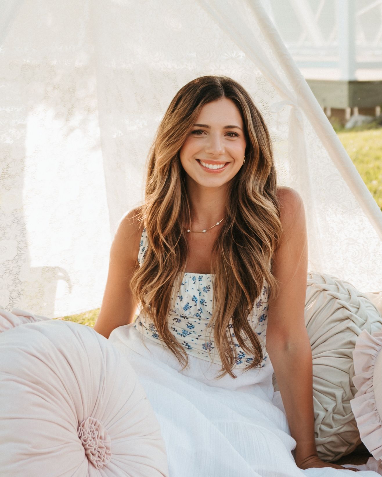 Smiling young woman with long wavy brunette hair sits among pastel pillows under a white lace canopy outdoors.