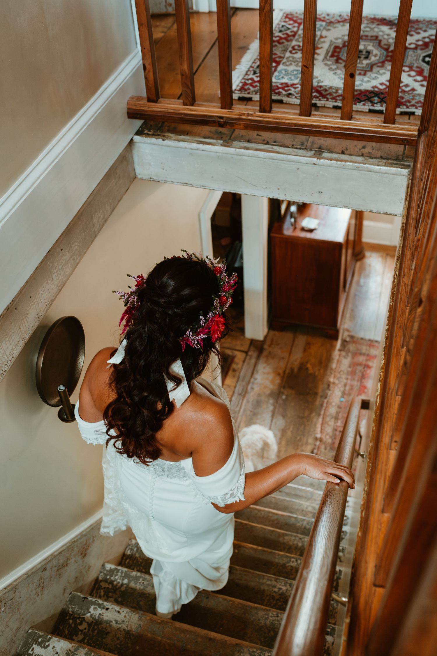 A person in a white dress descends a wooden staircase in an elegant indoor setting.